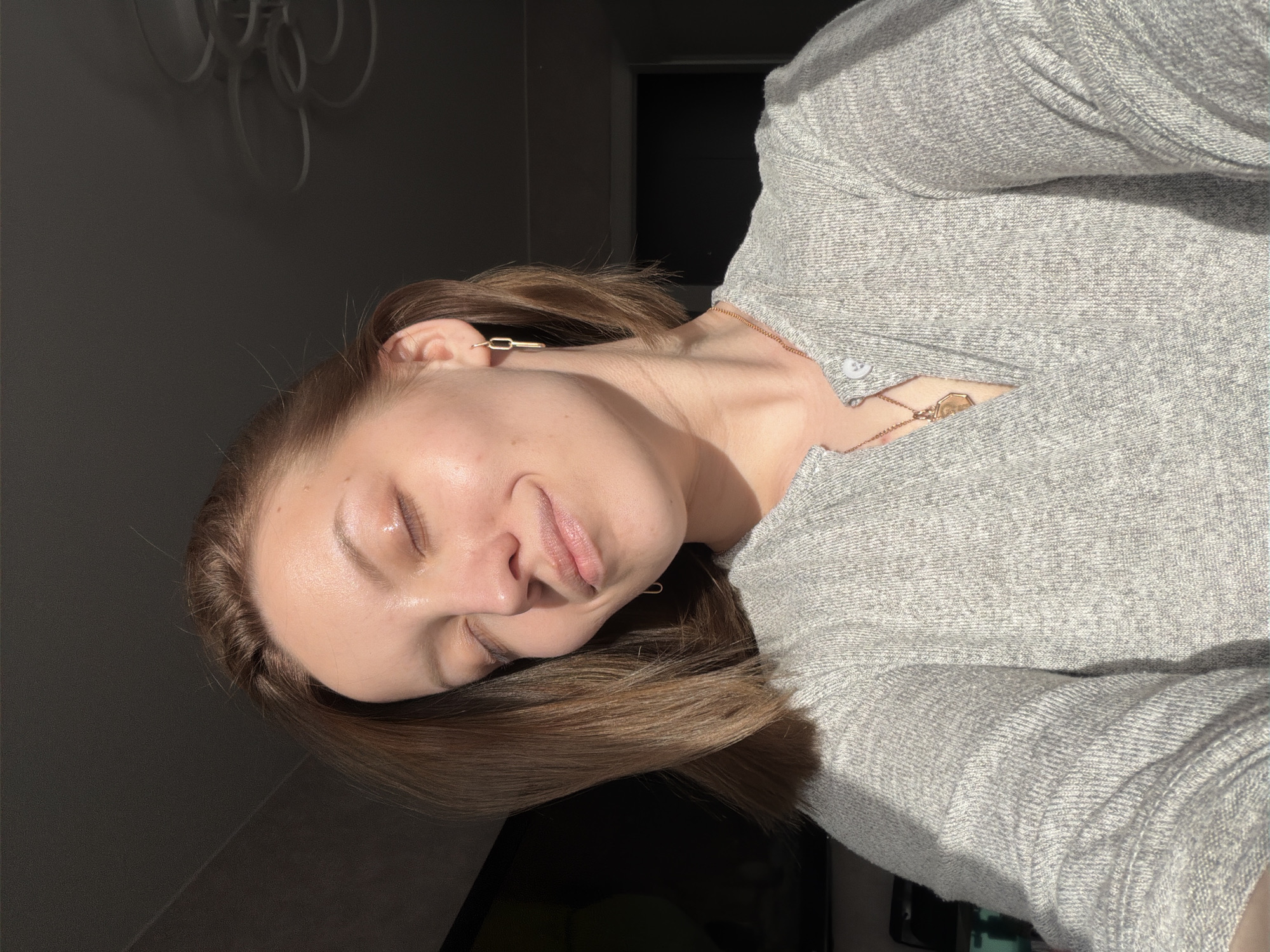 The image displays a woman's upper body and head, oriented sideways, appearing to be resting or reclining indoors. She has light brown hair and appears to be in her 20s or 30s. Her eyes are gently closed, and a soft, subtle smile graces her lips, conveying a sense of peace or contentment as she basks in bright, direct sunlight. The light strongly illuminates the right side of her face and upper body, creating highlights and shadows that emphasize her features and a dewy complexion.
She is dressed in a light grey, textured long-sleeved top or sweater. Accessories include a modern gold-colored, chain-link earring visible on her left ear, and a delicate gold-toned necklace with a small, hexagonal pendant partially visible near her collarbone.
The background is predominantly dark, likely an interior wall. Above her head, a light-colored decorative wall accent, featuring overlapping circular shapes, is visible. A dark rectangular area, possibly a doorway or a turned-off screen, occupies the lower right portion of the image. The strong, direct sunlight suggests the time of day is either morning or afternoon. The scene is set indoors, within a home in Reshetnikovo, Russia. No visible text is present.