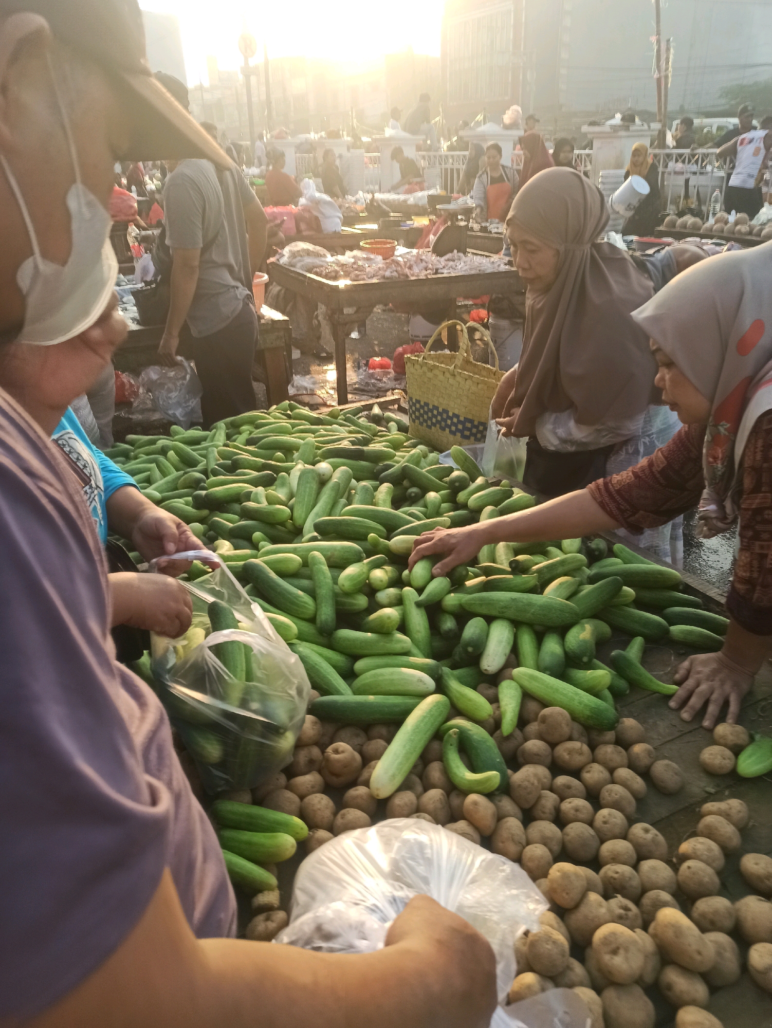This image depicts a busy outdoor market scene at sunrise in Cikarang, Indonesia. The foreground is dominated by a large pile of cucumbers and potatoes spread across wooden tables and the ground. Several individuals are visible, engaged in transactions or preparing produce. A woman wearing a brown hijab is seen sorting cucumbers, while another woman in a grey hijab is interacting with a customer near a basket. In the left foreground, a person in a purple shirt is holding a plastic bag with cucumbers, seemingly making a purchase. Other market-goers and vendors are present in the background, amidst stalls and various goods. The atmosphere is characterized by the soft glow of the early morning sun, casting a warm light over the bustling market.
