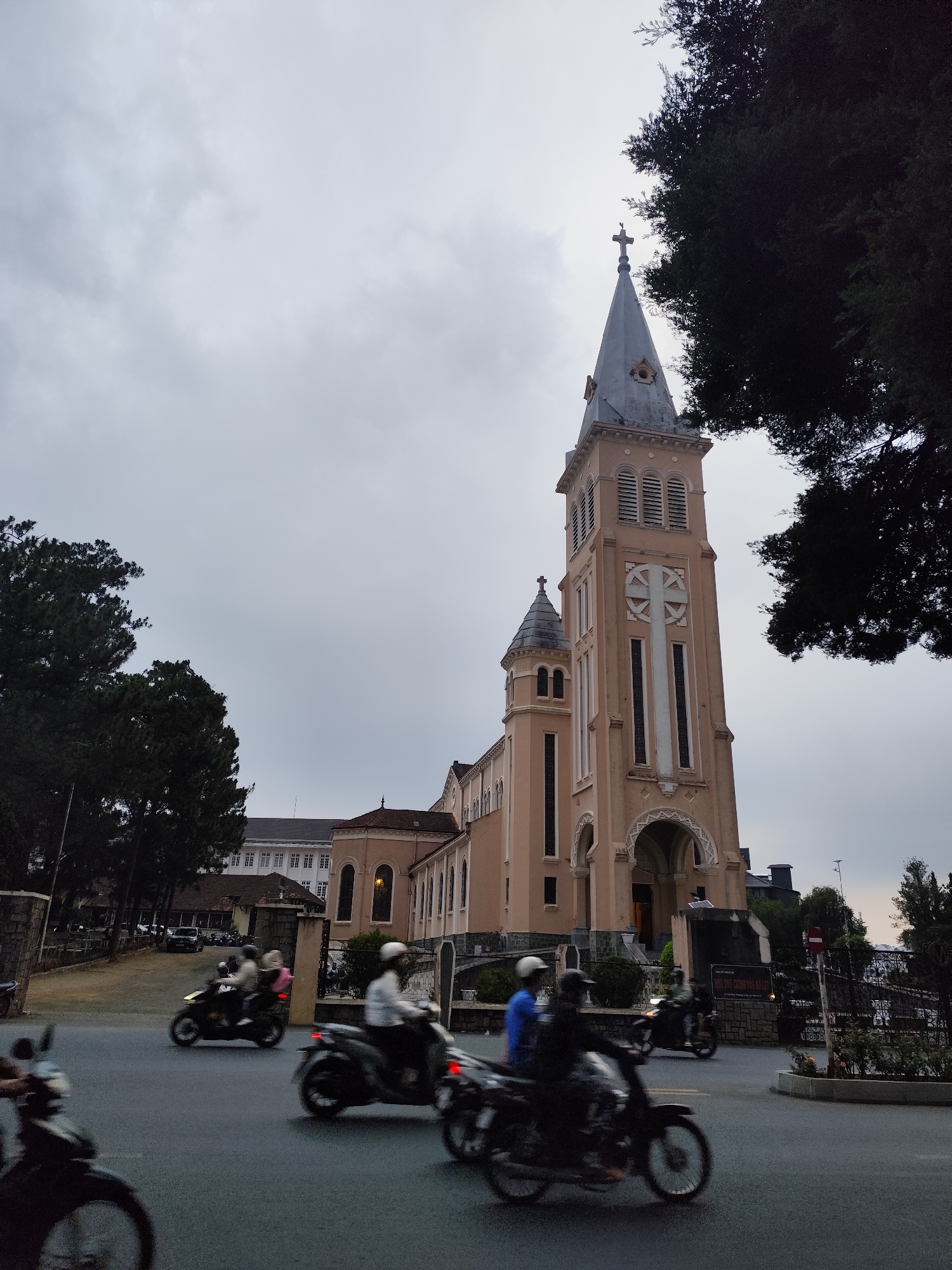 The image presents a vibrant street scene centered around the Da Lat Cathedral (Nhà thờ Con Gà) in Da Lat, Vietnam, captured on an overcast or late afternoon day. The sky is a uniform, muted gray, suggesting diffused light without direct sunlight.
The main subject is the large, light peach-colored Da Lat Cathedral, a prominent example of Gothic Revival architecture. Its most striking feature is the tall, elegant spire, crowned with a cross and a small rooster weather vane. The cathedral's main facade displays a large, white, stylized cross pattern and features several arched windows and entranceways. A smaller, secondary tower with a conical roof is visible on the left side of the main structure. Dense, dark green conifer trees frame the church on both sides, their foliage contrasting with the lighter tones of the building and the sky.
In the foreground and midground, a paved street is bustling with activity. Multiple motorbikes are depicted, many with motion blur, indicating they are in motion. Riders, wearing helmets, navigate the road. To the right, a red circular "no entry" sign stands near a decorative metal fence and a stone wall that separates the church grounds from the street. Behind the cathedral, other buildings are visible, including a multi-story structure with numerous white-framed windows.
Text on a sign near the church entrance is partially visible, stating "GIÁO XỨ CHÍNH TOÀ ĐÀ LẠT" and "NHÀ THỜ CHÍNH TÒA ĐÀ LẠT," which translates to "Da Lat Cathedral Parish" and "Da Lat Cathedral." The photograph captures a blend of historical architecture and the dynamic urban environment of Da Lat.