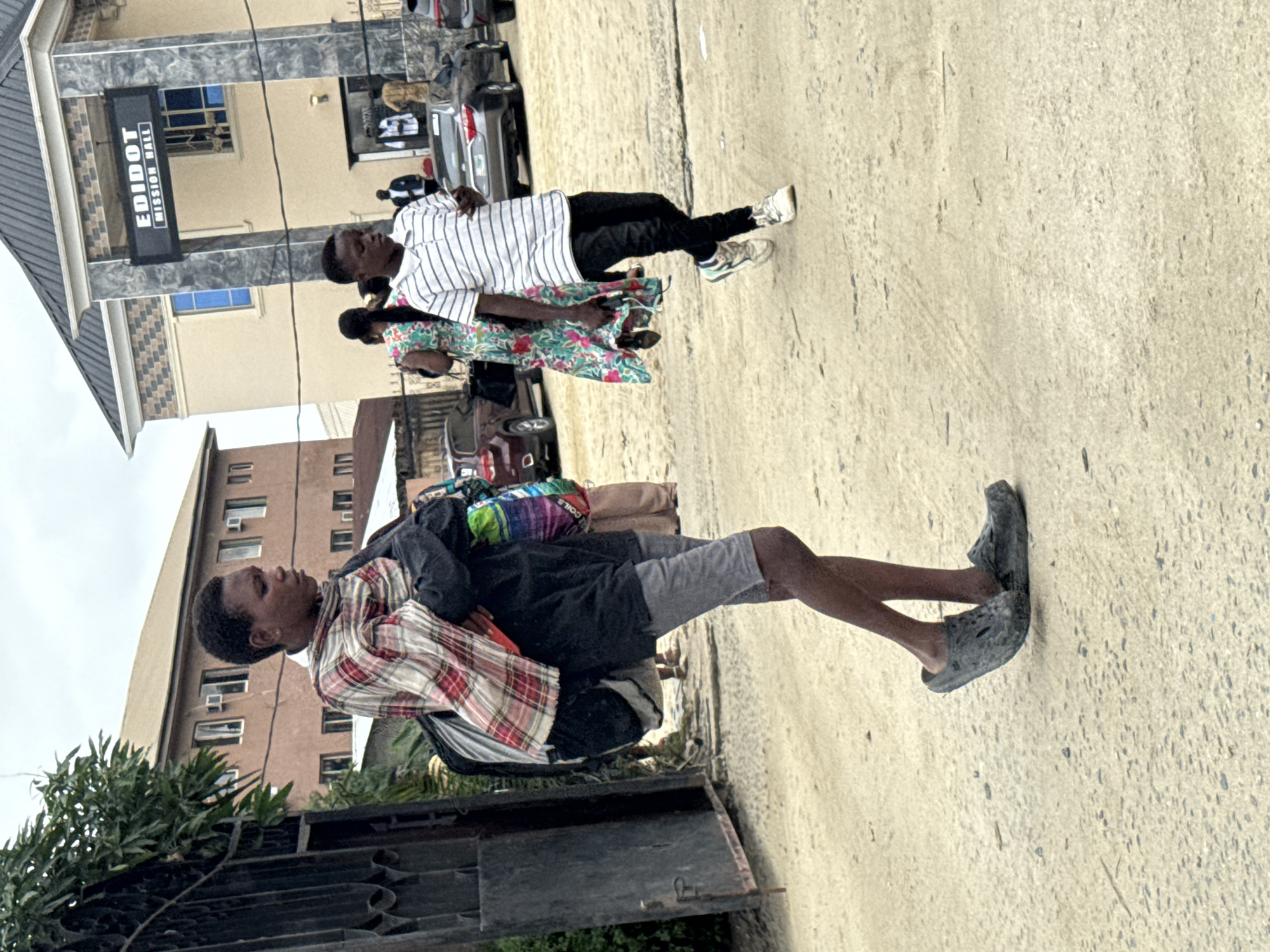 The image depicts a street scene in Ado, Nigeria, likely near a commercial or religious establishment. In the foreground, a young person walks forward, carrying a backpack and draped in a plaid shawl. They are wearing grey shorts and dark clogs. Behind them, another young person walks in the opposite direction, holding a phone and wearing a striped t-shirt, black pants, and white sneakers. In the background, several buildings are visible, including a prominent structure with a sign that reads "EDIDOT MISSION HALL." Other individuals and vehicles are present, suggesting a bustling urban environment. The ground is a dirt or sandy surface.