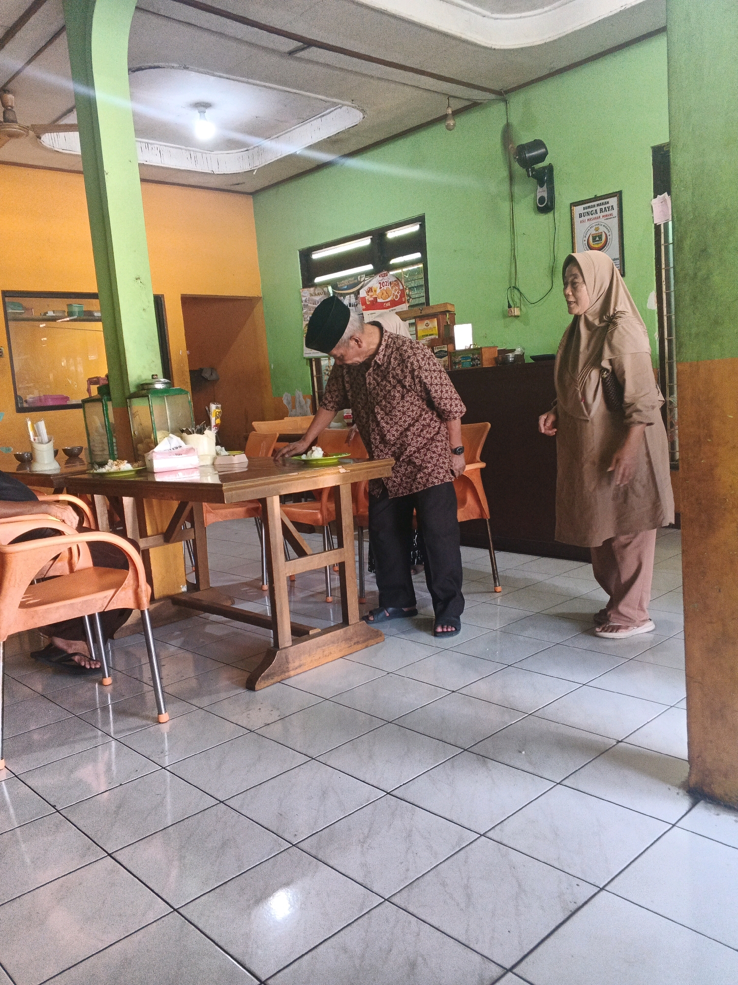 This image depicts the interior of a restaurant in Ungaran, Indonesia. Two individuals are present: an elderly man wearing a black peci hat and a patterned brown shirt, and a woman in a beige hijab and matching clothing. The man is leaning over a wooden dining table, appearing to be arranging food on a plate. The woman stands to his right, looking towards him. The environment is a simple dining area with green and orange walls. A wooden table with orange chairs occupies the center of the frame. Other visible elements include a ceiling fan, a wall-mounted fan, a framed sign with Indonesian text, and various food service items on the table, such as plates, napkins, and utensils. The floor is tiled in a white diamond pattern.