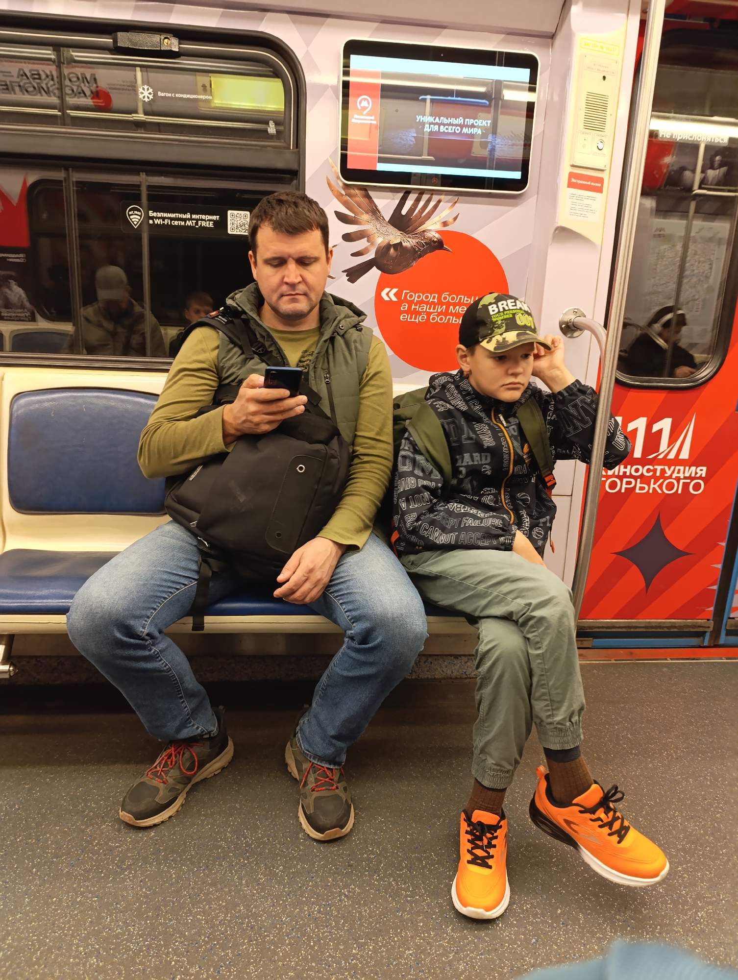 Two males, an adult and a boy, are seated on blue upholstered seats inside what appears to be a subway car, likely in Russia, given the visible Russian text. The man, on the left, wears a green long-sleeved shirt, a dark green vest, and blue jeans, with dark sneakers featuring red laces. He is holding a smartphone and looking down at it, appearing engrossed. A black messenger bag is slung across his body.
Beside him, to the right, sits a boy wearing a dark camouflage-patterned baseball cap, a black and white patterned hoodie, and light green cargo pants, with bright orange and black athletic shoes. He rests his left elbow on the armrest and his hand on his head, looking forward with a somewhat pensive or bored expression. A backpack is visible on his back.
The interior of the subway car features large windows reflecting other passengers, and white and grey patterned walls. Advertisements and informational signs are visible. A digital screen above the man's head displays "УНИКАЛЬНЫЙ ПРОЕКТ ДЛЯ ВСЕГО МИРА" (UNIQUE PROJECT FOR THE WHOLE WORLD). Another sign near the window states "Вагон с кондиционером" (Car with air conditioning). Below it, a sticker indicates "Безлимитный интернет в Wi-Fi сети MT_FREE" (Unlimited internet in Wi-Fi network MT_FREE) with a QR code. A large red advertisement on the right side features the number "111" and "КИНОСТУДИЯ ГОРЬКОГО" (GORKY FILM STUDIO). Another sign says "Не прислоняться" (Do not lean). The boy's cap has text partially visible as "BREAK YOUR PERSO". The lighting inside is artificial.