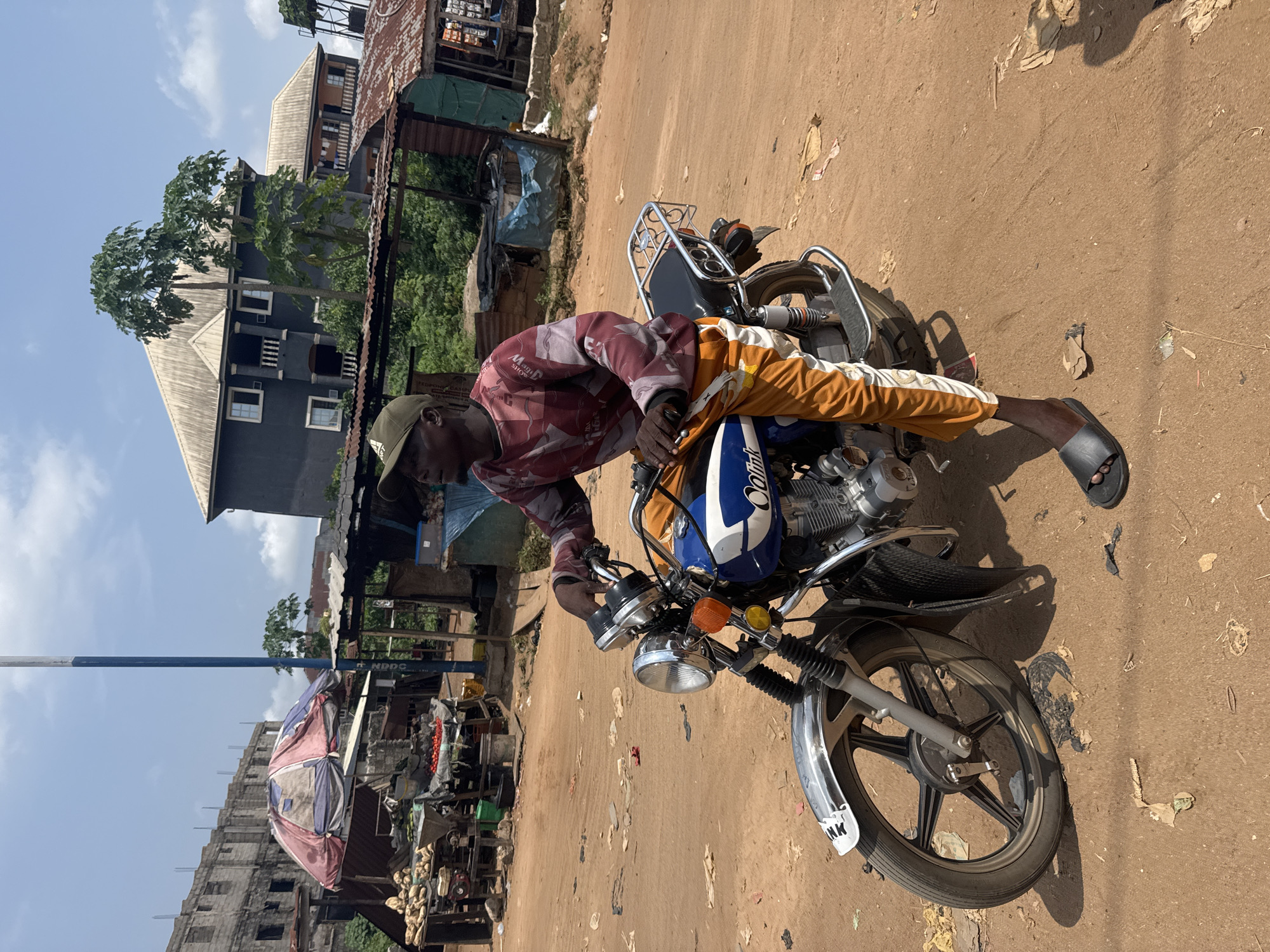 The image captures a daytime scene on an unpaved, reddish-brown dirt road in Ihiagwa, Nigeria. The sky is clear blue with scattered white clouds, and bright sunlight casts distinct shadows.
The central subject is a dark-skinned man, seen from the waist up, wearing a red and white patterned long-sleeved shirt, orange and white striped pants, and a green baseball cap. He has black sandals on his feet. The man is positioned beside a motorcycle, leaning over its handlebars, suggesting he is either preparing to ride, dismounting, or making an adjustment to the vehicle.
The motorcycle is primarily blue and white with chrome accents on its handlebars, front fender, and exhaust. It features a single headlight and orange turn signals. The front fender bears the partial text "NK" in white.
In the background, to the left, a tall blue and white pole stands next to a rudimentary market stall or roadside setup. This stall includes a colorful umbrella (red, white, and blue/grey), and displays produce such as yams and what appear to be tomatoes in sacks. An unfinished concrete building is also visible behind the market stall. Further into the background, there's a prominent multi-story building, dark grey/black with white window frames and a light-colored roof, surrounded by green trees. To the right, more makeshift structures with corrugated iron roofs and green foliage are visible. Scattered debris and paper litter the dirt ground. A sign in the background near the market stall shows partially visible text, including "NPDC".