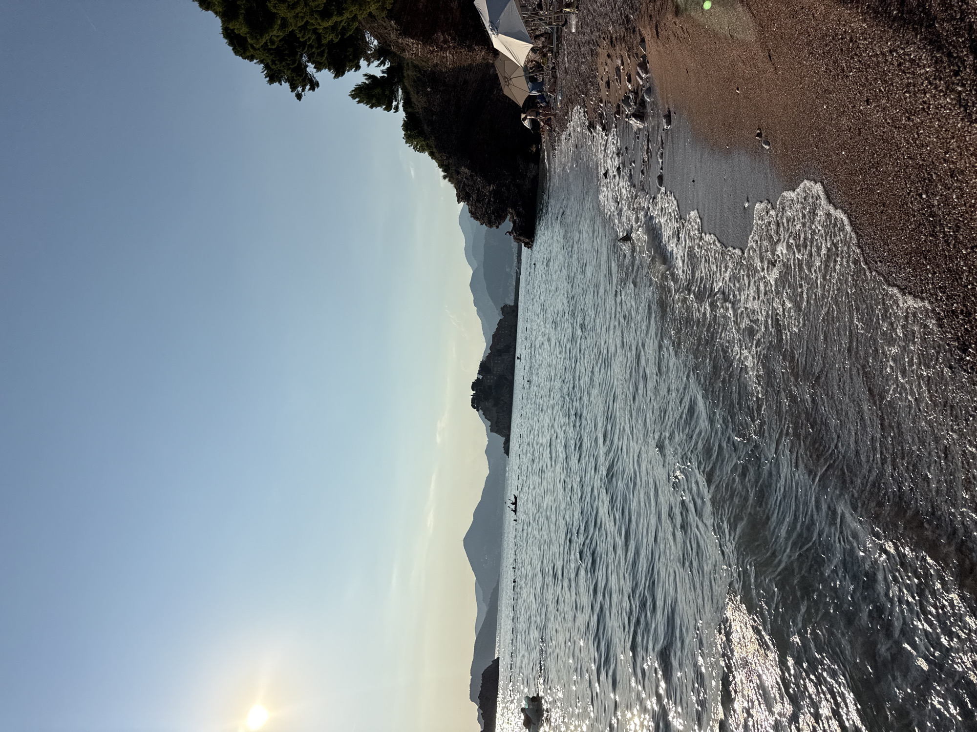 This is a vertical, landscape-oriented photograph of a beach and the sea. The image is dominated by the bright, clear sky and the shimmering water. On the left side of the frame, the sun is visible low in the sky, casting a warm glow. Above the water, on the right side of the frame, a dark, rocky cliffside is covered with lush green trees.
Along the shoreline, a narrow strip of dark sand is visible, littered with small stones and pebbles. Waves are gently breaking on the sand, creating a foamy white edge. In the distance, silhouetted against the hazy sky, are layers of mountains, suggesting a coastal mountain range. There appear to be a few small figures in the water in the mid-ground, possibly swimming or engaged in water activities. A beach umbrella and some beach chairs are visible on the sandy area, indicating a popular spot.
The overall mood of the image is serene and peaceful, capturing a beautiful summer day at the beach. The light suggests it is late afternoon or early evening, with the sun beginning its descent. The scene is likely a tourist location, given the presence of beach amenities and the picturesque natural scenery. No visible text is present in the image.