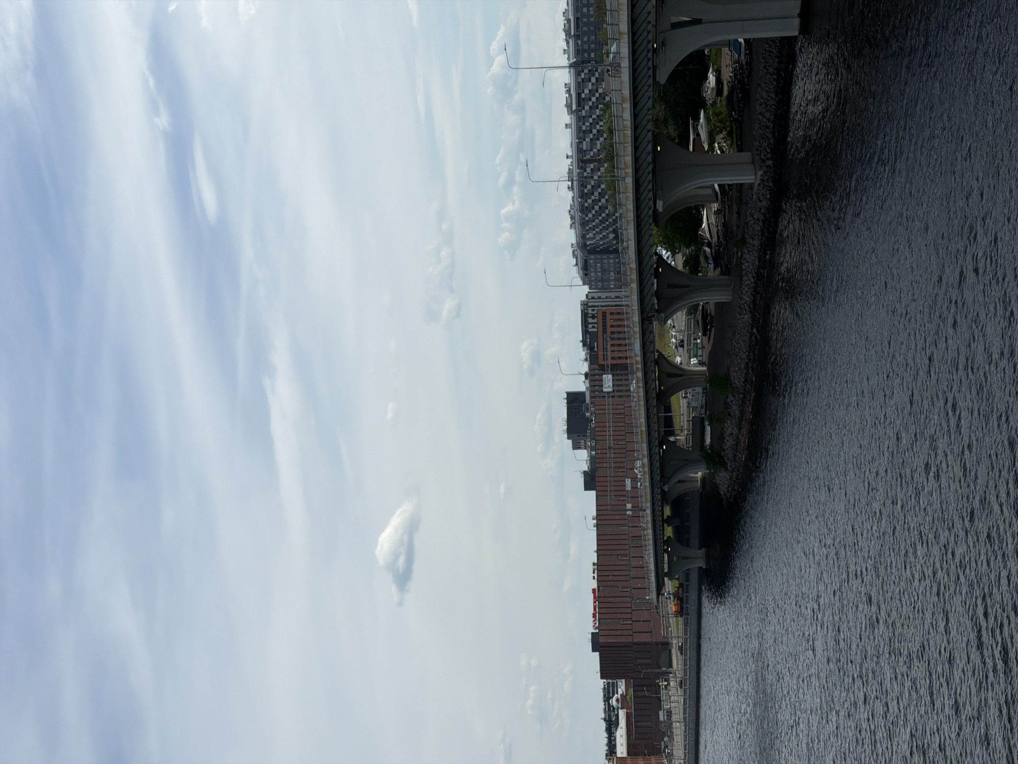 The image depicts an urban waterscape in Saint Petersburg, Russia, under a light blue sky with scattered cirrocumulus and stratocumulus clouds. In the foreground, a dark, rippled body of water, likely a canal, extends across the lower half of the frame.
A multi-span concrete bridge, part of a highway, traverses the scene horizontally. Its structure consists of several light-grey concrete piers and arched supports. The bridge carries a multi-lane roadway, on which several vehicles are visible, and is equipped with numerous streetlights featuring curved poles.
On the left side, behind the bridge, stands a large, multi-story building constructed primarily of dark red brick, with a flat roof. A prominent red sign with white lettering displaying "Honeywell" is mounted on its roofline. Further left, partially obscured, are other industrial or commercial structures and a parked orange construction vehicle near the water's edge.
To the right, beyond the bridge, a modern, multi-story building with a distinctive checkered facade of alternating dark and light square panels is visible. Further in the background, additional buildings with varying architectural styles and dark rooftops are present. Beneath the right section of the bridge, a rocky shoreline is exposed, where multiple small boats and boat trailers are stored on the land. No specific individuals are discernible in the scene. The lighting suggests daytime conditions with diffuse sunlight.
This scene is located in the Krasnoselsky District of Saint Petersburg. The prominent bridge is a section of the Western High-Speed Diameter (ZSD) highway, crossing either the Matisov Canal or the Duderhof Canal. The distinctive checkered building is part of the "Baltic Pearl" (Балтийская Жемчужина) residential complex. The large brick building with the "Honeywell" sign is situated in a commercial or industrial area adjacent to the highway, likely near Peterhofskoye Shosse.