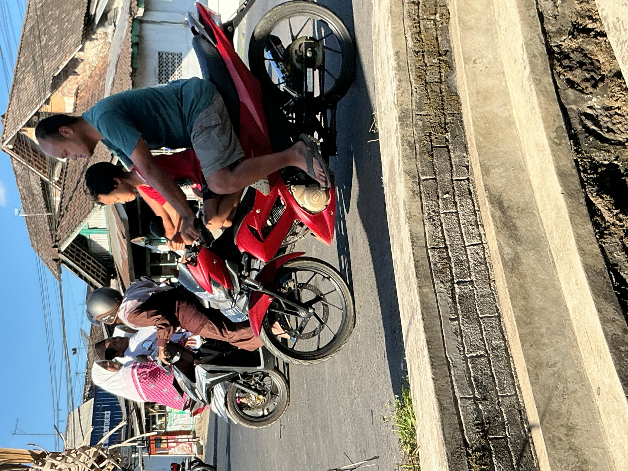 A man and a child are riding a red motorcycle in Kebonrejo, Indonesia. The man, wearing a teal t-shirt and grey shorts, is driving. The child, wearing a red shirt, is sitting in front of him. Both are looking to the right. Behind them, another motorcycle is carrying three people: a woman in a pink and white headscarf and patterned top, a man wearing a white shirt and black cap, and a person in a brown tunic and headscarf. The motorcycles are on a paved road. In the background, there are buildings with tiled roofs and utility poles. The foreground shows a concrete structure with a brick wall and steps. The sky is clear and blue.