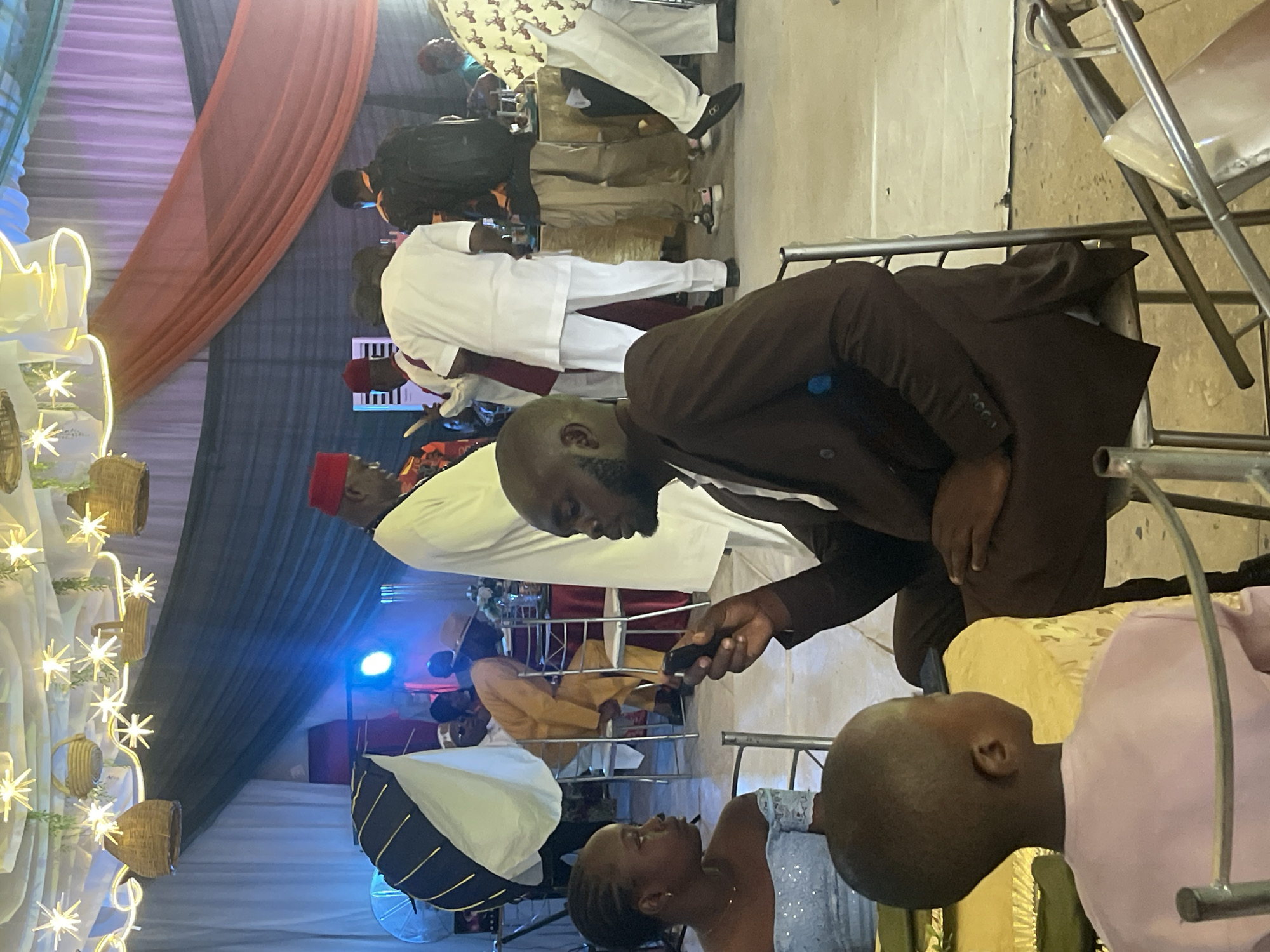The image shows an indoor event, possibly a wedding reception, in Jos, Nigeria. Multiple people are present, including a man in a brown suit holding a phone, a woman in a shimmering dress looking up at him, and other attendees in traditional attire and Western clothing. Seating includes metal chairs. There are decorative elements such as string lights and fabric drapes. The lighting includes spotlights and what appears to be a photography softbox. The overall setting suggests a celebratory atmosphere within a decorated venue.