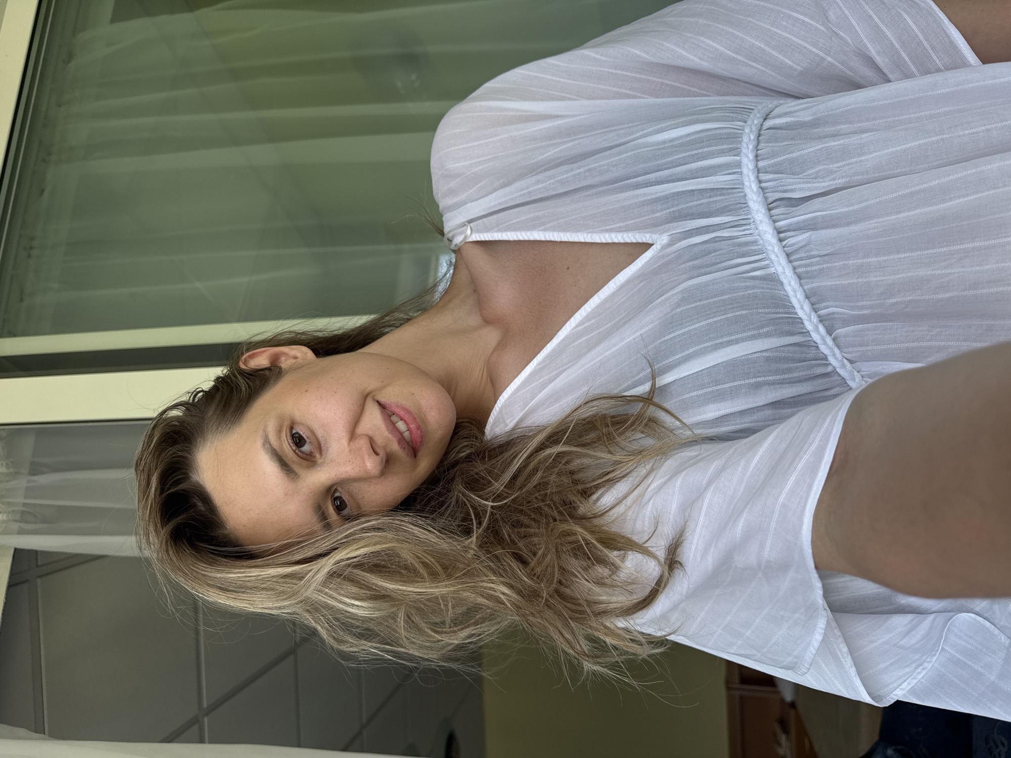A blonde woman is taking a selfie in front of a window with sheer white curtains. She is wearing a white, translucent dress with a V-neck and braided detailing at the bust. Her hair is styled in loose waves, and she is smiling. The background appears to be indoors, possibly a hotel room, with furniture visible in the distance. The scene is well-lit, suggesting natural light. The location is Nebug, Russia.
