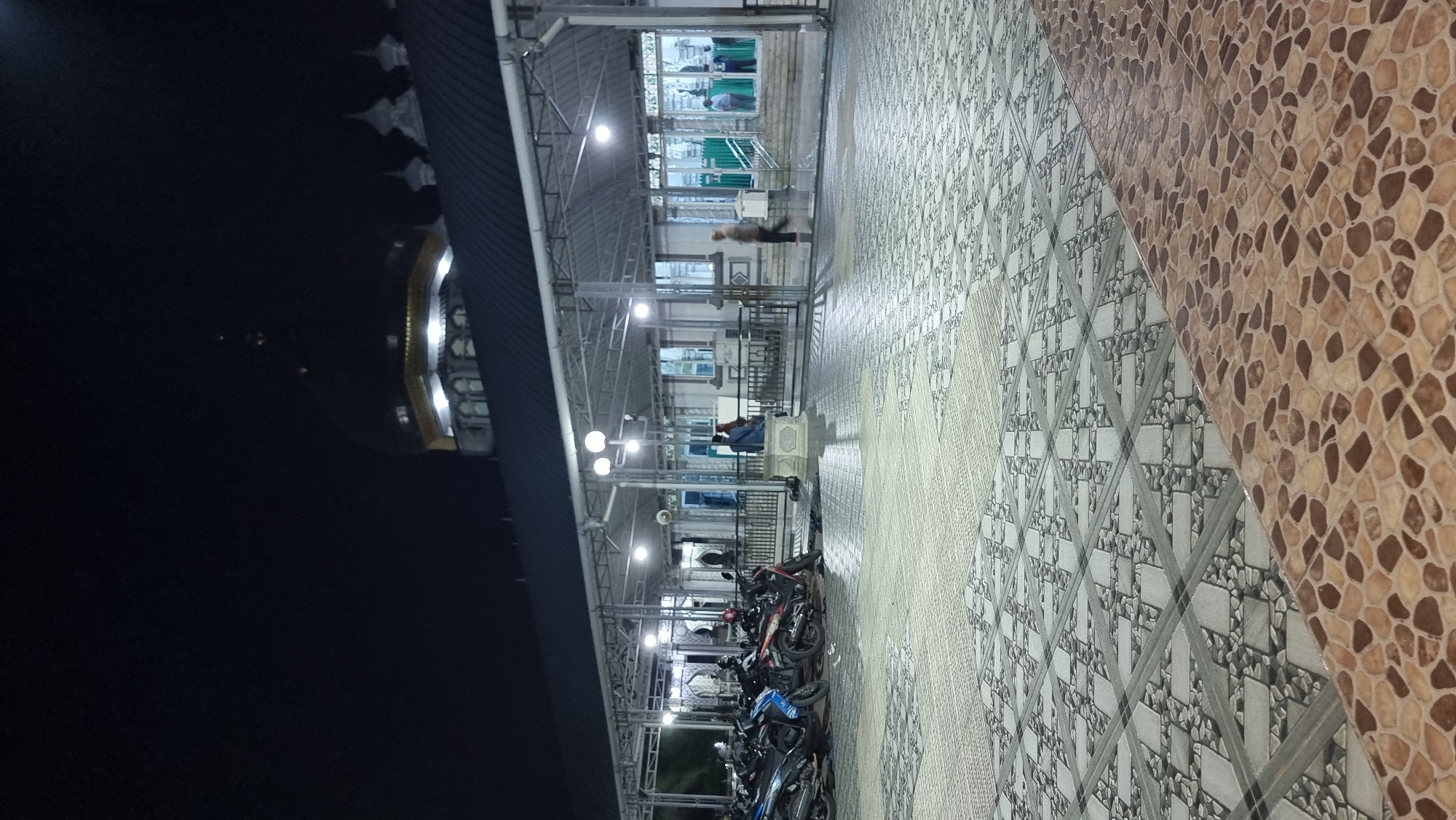 The image depicts the exterior of a mosque at night in Keudekruenggeukueh, Indonesia. The foreground is dominated by a tiled courtyard. To the left, a row of parked motorcycles is visible under a covered walkway supported by metal beams and pillars. The walkway extends to the right, revealing people standing and walking near the mosque entrance. The mosque itself features a large, dark dome with a golden band and illuminated windows. Other architectural elements of the mosque, including smaller domes and arches, are visible against the dark night sky. Several lampposts are illuminated, casting light on the courtyard and walkway. The overall impression is one of a serene and illuminated religious site at night.