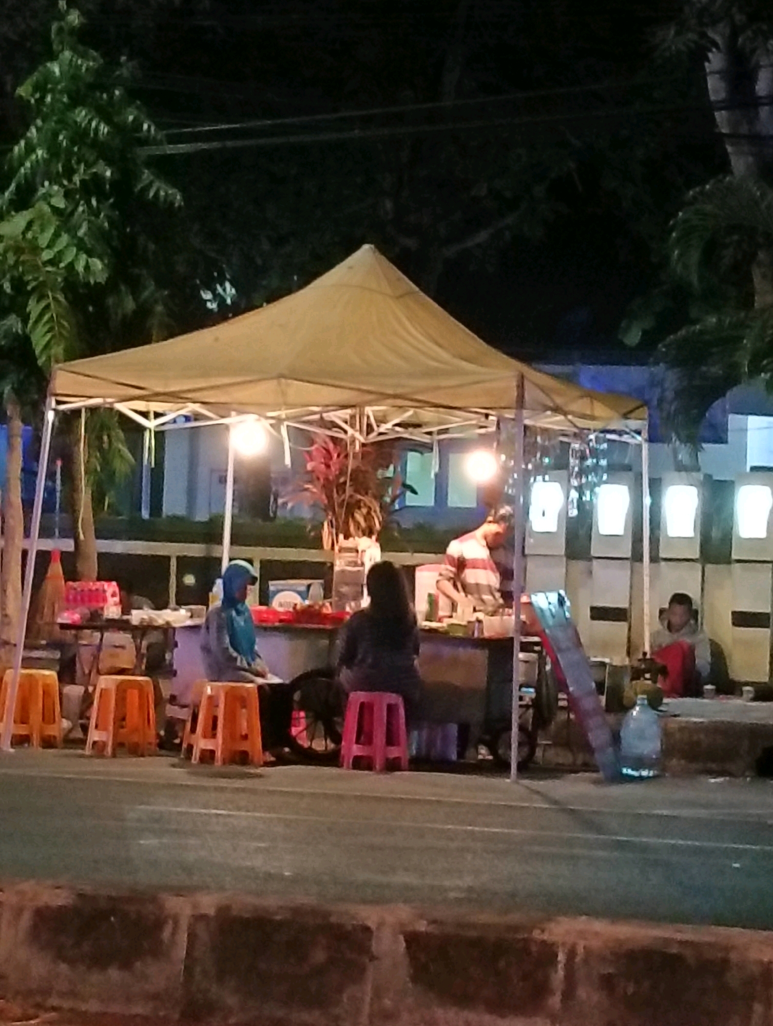 A nighttime outdoor scene shows a food stall set up under a yellow awning. Several people are visible, some seated on orange and pink plastic stools in front of the stall, and others standing behind it. The stall is illuminated by a bright lightbulb, and there are various items displayed on tables, including drinks and food. Behind the stall, a building with several windows is visible. The ground in front of the stall appears to be asphalt, with a low concrete barrier in the foreground. The overall impression is a busy street food vendor operating in an urban environment. The scene is located in Purworejo, Indonesia.