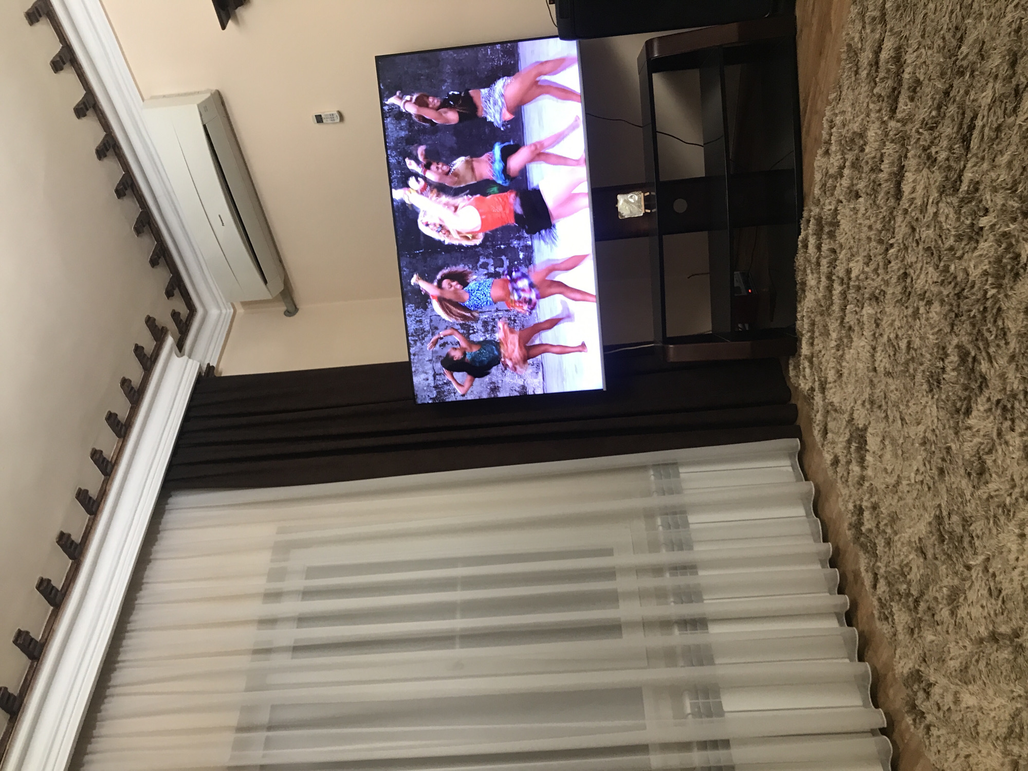 The image shows a living room with a television mounted on the wall, playing a scene of people dancing. Beneath the television, a window is covered with translucent blinds and dark brown curtains. To the right, a dark wooden entertainment center stands beside a beige shaggy rug. Above the television is an air conditioning unit. The room has beige walls with decorative trim. While it is not possible to pinpoint the exact location, the image is stated to be in Yerevan, Armenia.