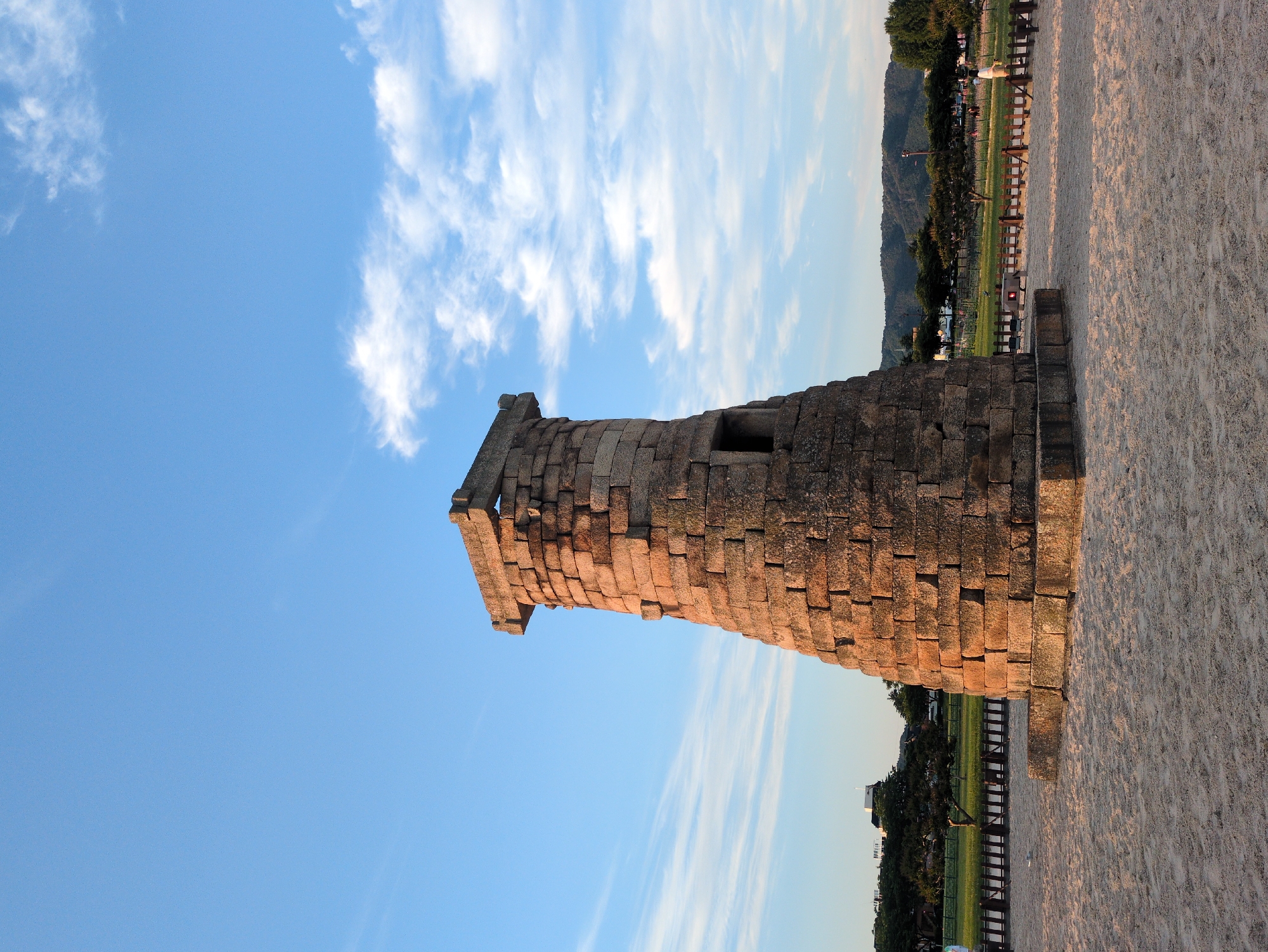 The image shows the Cheomseongdae observatory, a historic astronomical observatory located in Gyeongju-si, South Korea. The structure is a cylindrical stone tower composed of numerous rectangular stones. It has a window-like opening near the middle. A barrier separates the tower from a sandy surface. Beyond the barrier is greenery, trees, and distant buildings. The sky is blue with wispy clouds. The image appears to be rotated 90 degrees clockwise.