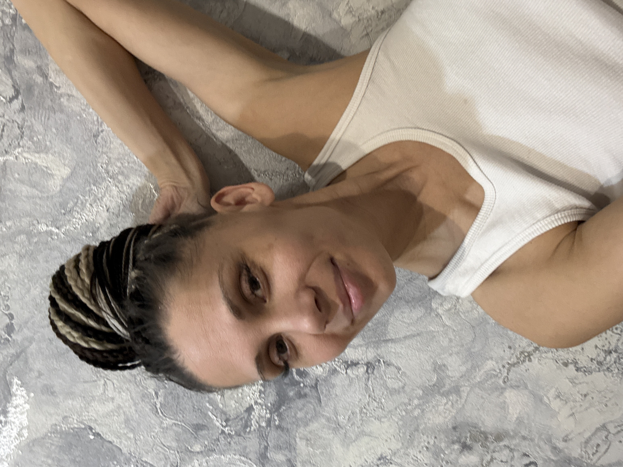 A woman is depicted lying down, oriented sideways with her head turned towards the viewer. She wears a plain white ribbed tank top. Her arms are extended upwards, resting on the textured surface behind her. Her hair is styled in a bun, composed of multi-toned box braids in black, brown, and lighter blonde/brown hues. She has a gentle, closed-mouth smile and looks directly at the camera with a calm expression. The background is a distinctively textured wall or surface, featuring a mottled pattern in shades of gray and white. The lighting is soft and appears to be from an indoor source, with no strong shadows. No specific time of day or weather is discernible beyond the indoor setting. There is no visible text in the image. The scene is located in Rusanovo, Russia.