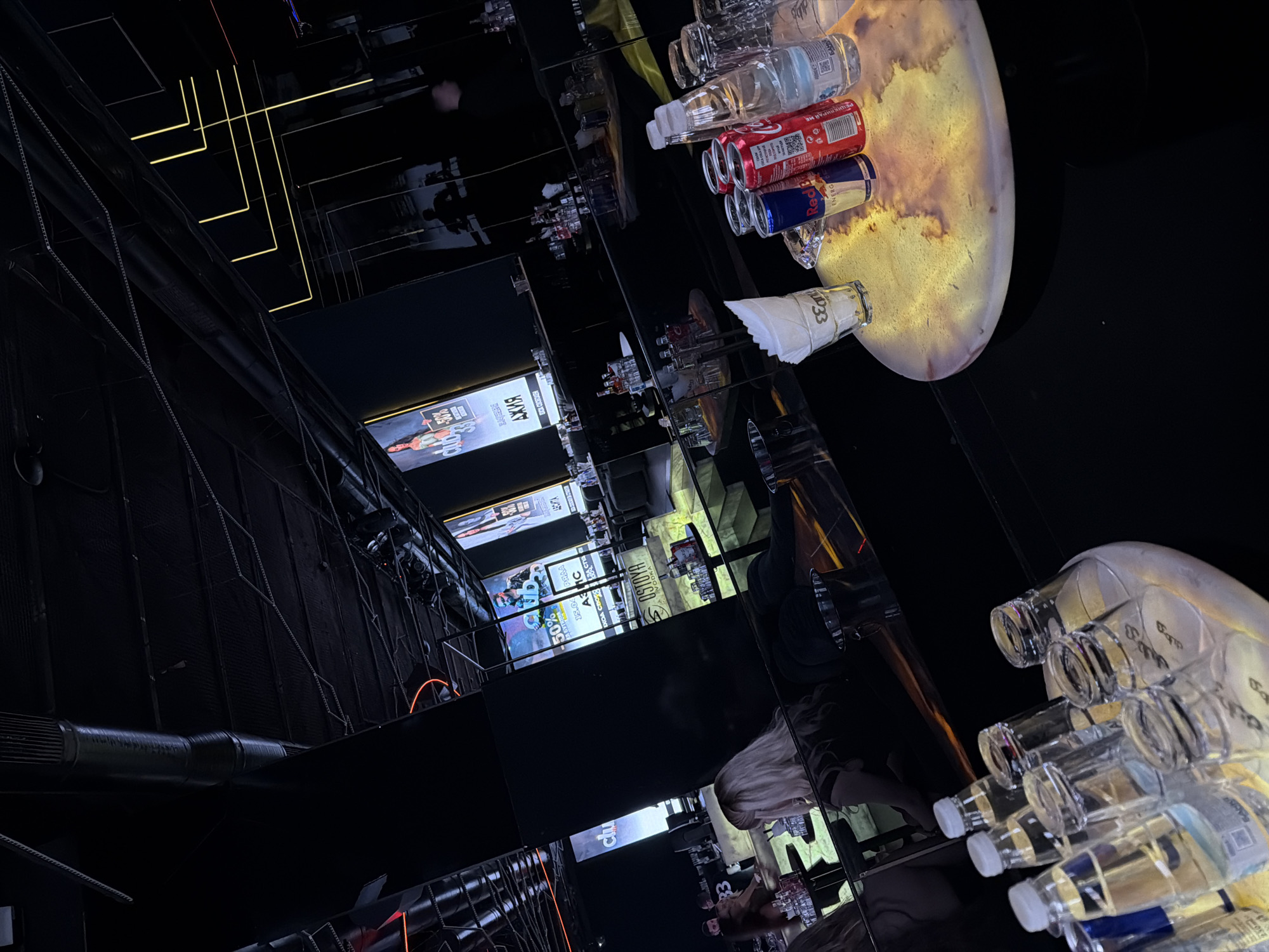 The image shows the interior of a bar in Sofia, Bulgaria, reflecting off of mirrored surfaces. There are bottles of water, cans of Coca-Cola and Red Bull, and glasses on a table with a light beneath it. Posters with advertisements are visible in the background, along with lighting fixtures on the ceiling. There are people reflected in the mirrors around the bar. The overall aesthetic is dark with bright, focused lighting.