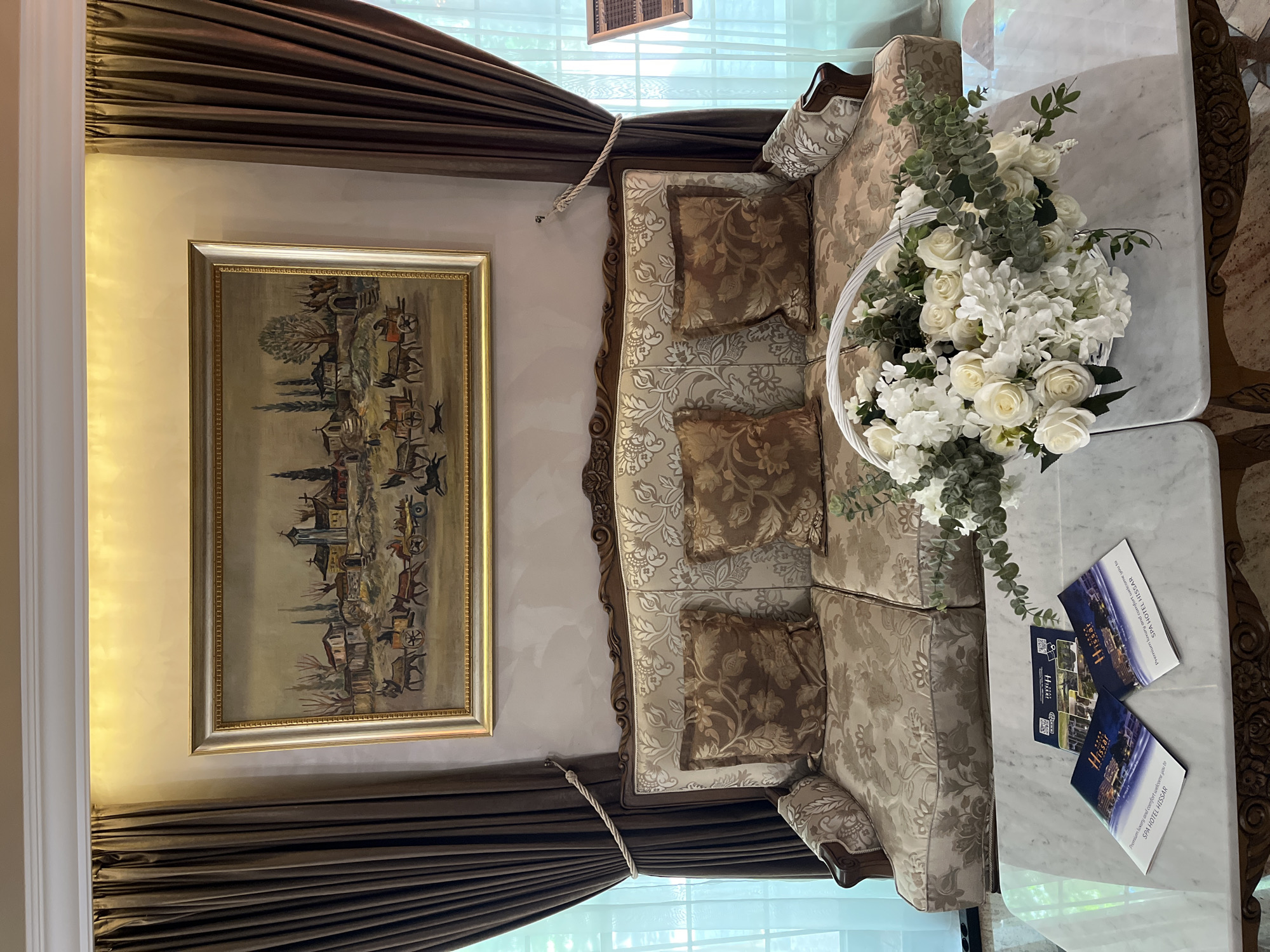 This indoor shot shows a room with beige and brown tones, featuring a painting, a sofa, and decorative objects. The painting, framed in gold, depicts a village scene and hangs on a wall with brown draped curtains on either side. Beneath the painting, a three-seater sofa is positioned, upholstered in a beige and brown floral pattern with matching cushions. A bouquet of white flowers is placed on a marble-topped side table next to the sofa. Several promotional leaflets with the word "Hissar" and "SPA HOTEL HISSAR" printed on them are also on the table. The room's location is not evident from the image alone, but the pamphlets suggest a connection to the "Spa Hotel Hissar." The picture is taken from an elevated angle, focusing on the furniture and decoration.