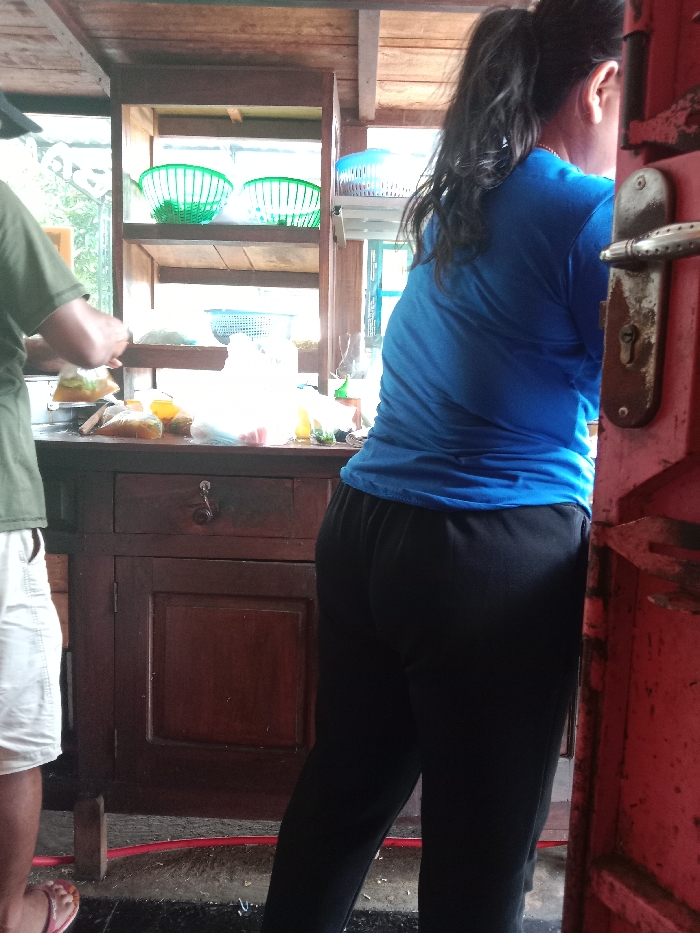 A woman with dark hair pulled back in a ponytail stands in black pants and a bright blue shirt. She faces away from the viewer. To her left, a person wearing a green shirt and white shorts stands at a brown wooden counter. The counter holds items including plastic bags and containers. A tiered shelf behind them holds green and blue baskets. A red doorframe is visible to the right of the woman, with a silver handle and lock.
