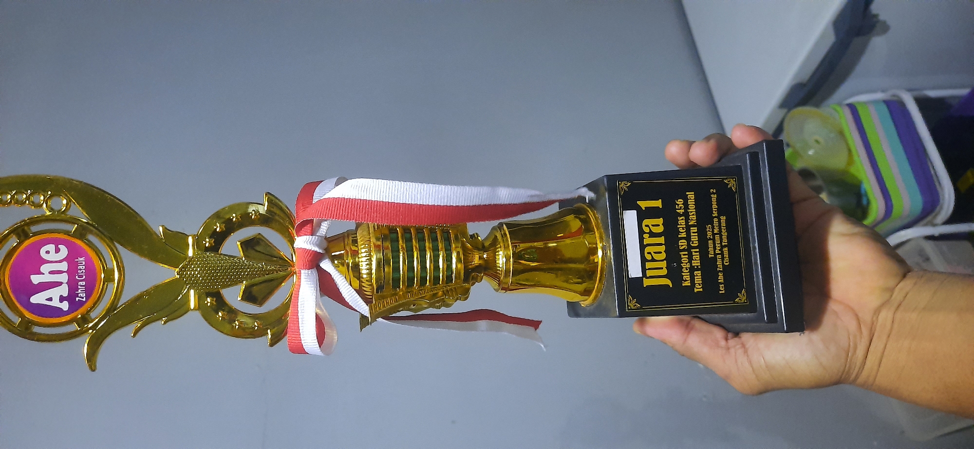 A person's hand is holding a golden trophy with a black base. The trophy has a decorative top with a circular emblem that reads "Ahe Zahra Cisauk". A red and white ribbon is tied around the trophy. The black base of the trophy has a plaque with text that reads "Juara 1", "Kategori SD kelas 456", "Tema: Hari Guru Nasional", "Tahun 2025", and "Les Ahe Zahra Perum Metro Serpong 2 Cisauk Tangerang". In the background, there is a blurred wall and a colorful container. The trophy appears to be a prize for a competition held in Cisauk, Tangerang, Indonesia, likely related to National Teacher's Day for elementary school students in grades 4-6.