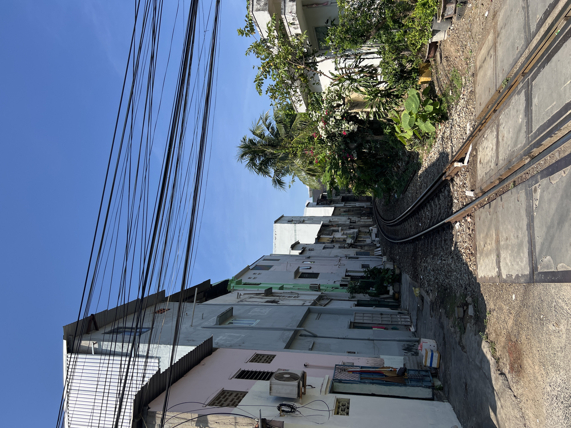 The image depicts a narrow railway track running between closely spaced buildings in what appears to be a residential area of Nha Trang Ward, Vietnam. The buildings are multi-story structures of varying colors, with visible signs of age and wear. A profusion of power lines stretches across the sky above the buildings, creating a dense network. The railway track is flanked by concrete blocks and gravel. A few trees and plants are visible along the track and near the buildings, adding a touch of greenery. The sky is clear and blue.