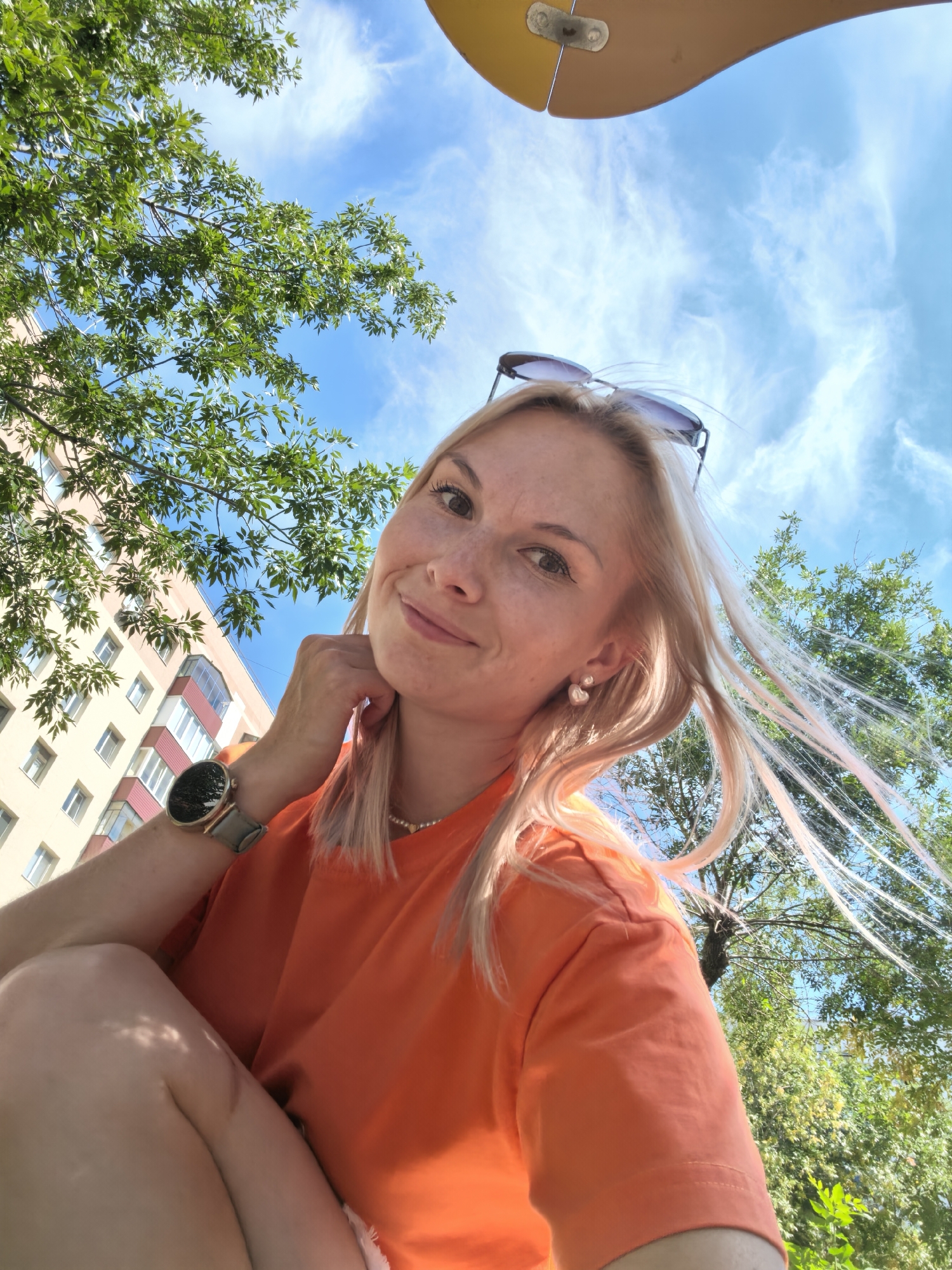 An outdoor, low-angle, selfie-style, medium shot shows a young, fair-skinned woman with blonde hair, freckles, and blue eyes. She is wearing an orange t-shirt, a pearl necklace, pearl earrings, sunglasses perched on her head, and a gray-banded watch on her left wrist. She is resting her chin on her hand and smiling. Her hair is blowing in the wind. In the background, a light brown apartment building with white-trimmed windows is visible, along with green trees and a blue sky with wispy clouds. The scene is set in Sterlitamak, Russia.
