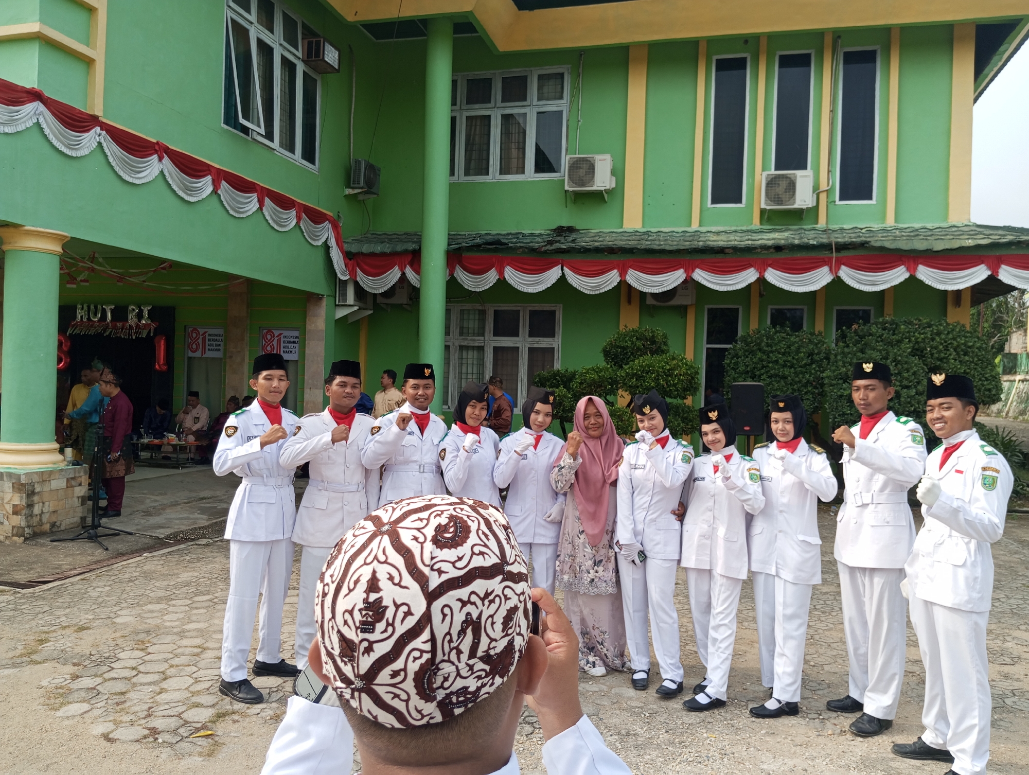 In Pematang Reba, Indonesia, a group of young people in white uniforms stand posed for a photograph in front of a bright green building adorned with red and white bunting. The building has several windows and an air conditioning unit visible. Most of the individuals are wearing black skullcaps and red scarves around their necks, with patches on their uniforms. One person in the foreground is wearing a traditional patterned cap and appears to be taking the photo. Other people are visible in the background, some in more traditional attire. The ground is paved with hexagonal tiles, and there are bushes and trees visible. The overall scene suggests a formal event or celebration.