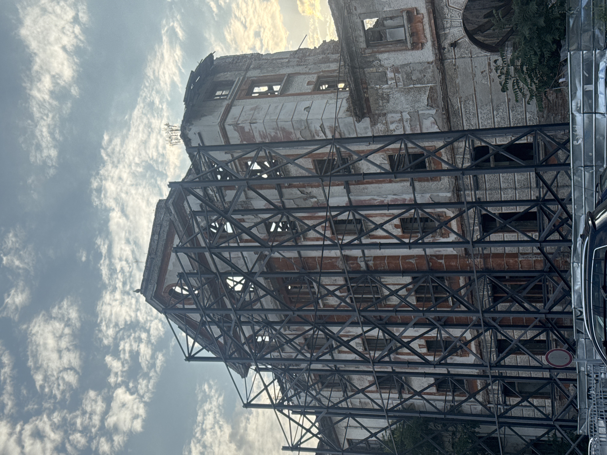 This image depicts a severely dilapidated industrial building, identified as part of the Royal Tobacco Factory complex in Plovdiv, Bulgaria. The multi-story structure exhibits extensive damage, with large sections of its pale exterior plaster having peeled away, revealing the underlying red brickwork. Numerous window openings are empty or contain broken frames, and the roofline is uneven and partially collapsed.
A substantial, dark metal scaffolding system, characterized by intricate X-bracing, envelops the central and left portions of the building's facade, indicating structural reinforcement, stabilization, or preparation for extensive renovation or deconstruction. On the highest intact point of the central roof, a single white bird, consistent with a seagull, is perched. To the right, another section of the building shows further advanced decay, including a large arched opening at ground level.
The sky is partly cloudy, with scattered white clouds against a light blue backdrop. Soft, diffuse sunlight appears to illuminate the upper right side of the building, suggesting either early morning or late afternoon. In the foreground, a low, corrugated metal construction fence runs horizontally, partially obscuring the roof of a dark-colored vehicle and a circular "No Entry" traffic sign positioned directly in front of the building. Green foliage from trees and shrubs is visible on both the left and right sides of the structure.