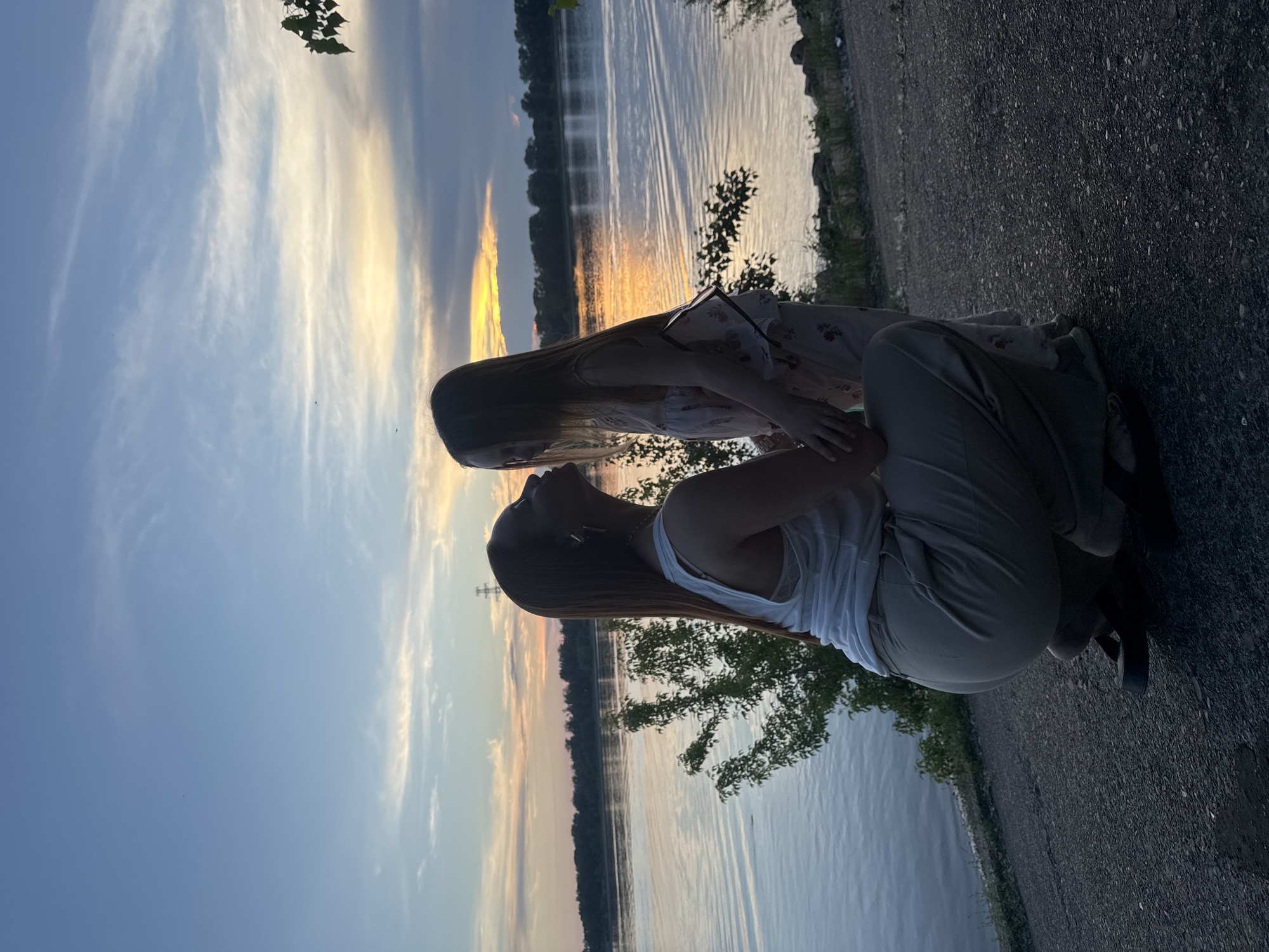 The image shows two people in silhouette by a body of water at sunset. One person is kneeling and the other is leaning forward, with their heads close together, possibly interacting. The sky is filled with wispy clouds, and the water reflects the warm colors of the sunset. The scene appears to be a peaceful outdoor setting, likely a park or recreational area along a river or lake. The time of day is evident from the golden hour lighting. The ground in the foreground is a gravel path, and there are some trees and rocks visible along the water's edge. The mood appears to be serene and intimate. There is no visible text in the image.