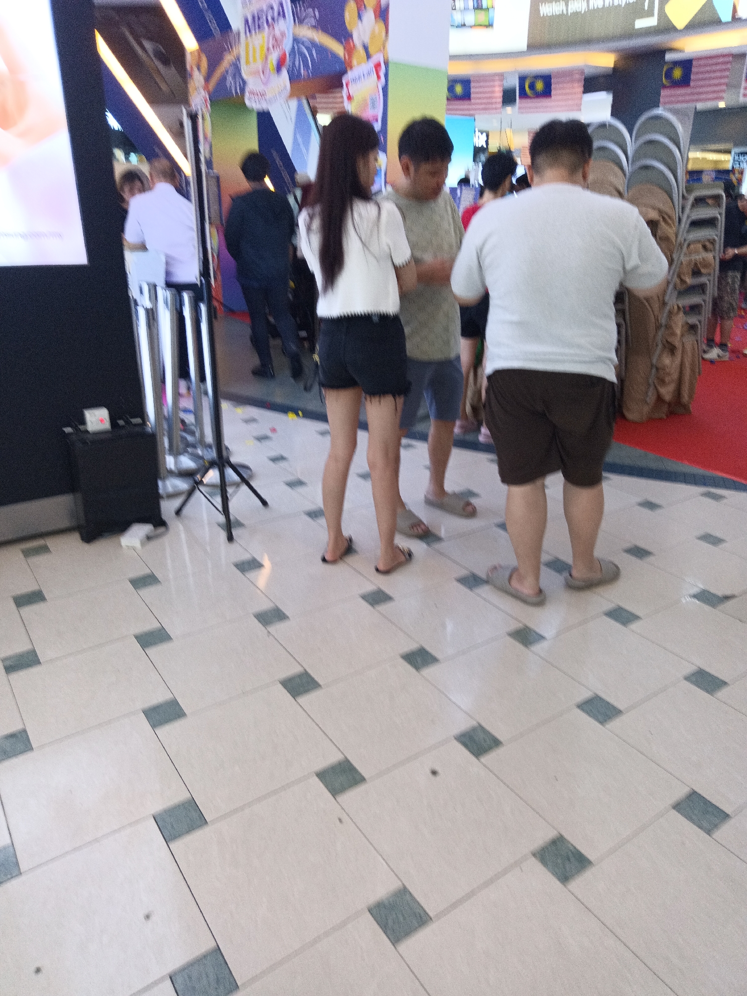 The image captures a scene inside a shopping mall, likely in Kuala Lumpur, Malaysia, given the Malaysian flags displayed prominently in the background. The floor is tiled with large, light-colored rectangular tiles interspersed with smaller, darker square tiles. Several people are visible, primarily from the waist down.
In the foreground, two individuals are standing close together. The person on the left is wearing a white t-shirt and dark shorts, with their back to the camera. They are wearing light-colored flip-flops. To their right, another person is wearing a light grey t-shirt and dark shorts, also with their back to the camera, and they are wearing light grey sandals.
Behind them, a woman in a white top and black shorts is facing away from the camera, her legs and feet visible. She is wearing dark flip-flops. To her left, a man in a dark outfit is walking away from the camera. Further back, a person in a white shirt is visible.
In the background, stacked chairs are visible, some covered with brown fabric. Banners, one with the word "MEGA" and what appears to be an illustration of a beverage, are hanging from above. The overall environment suggests a public indoor space, possibly an event or promotional area within the mall.