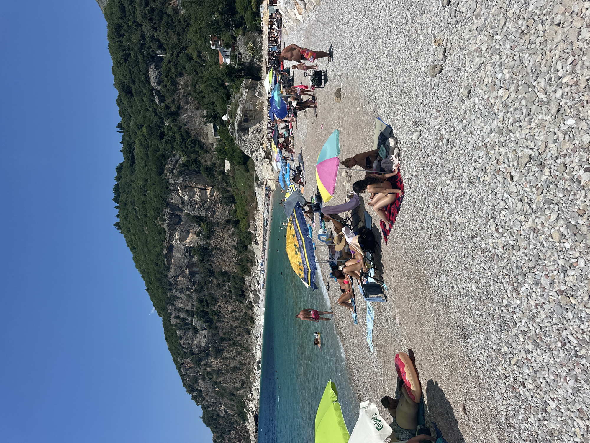 This media file captures a lively beach scene on a sunny day in Mirište, Montenegro. The image is oriented vertically, showing a crowded pebble beach bordering a clear, turquoise sea. The beach is dotted with numerous colorful beach umbrellas and people relaxing on towels and beach chairs.
The subjects are primarily people enjoying a summer day at the beach. Many are sunbathing, sitting under umbrellas, or wading in the shallow waters. Some individuals are visible swimming or enjoying the sea. Objects include a variety of beach umbrellas in shades of blue, yellow, pink, and green, as well as beach chairs, towels, and a distinctive inflatable ring shaped like a donut. The setting is a natural beach with a dramatic, forested cliff face rising steeply on the left side of the frame, and a clear blue sky overhead.
The activity depicted is typical of a beach day: recreation, relaxation, and enjoying the warm weather. The time of day appears to be midday, given the bright sunlight and distinct shadows. The weather is sunny and warm, ideal for beach activities. The location is identified as Mirište, Montenegro, with the mountainous terrain and the style of the beach providing visual cues. There are no strong emotions conveyed, but the general atmosphere appears relaxed and leisurely.
There is some visible text on a white t-shirt in the lower right portion of the image, which appears to read "Carlsberg".