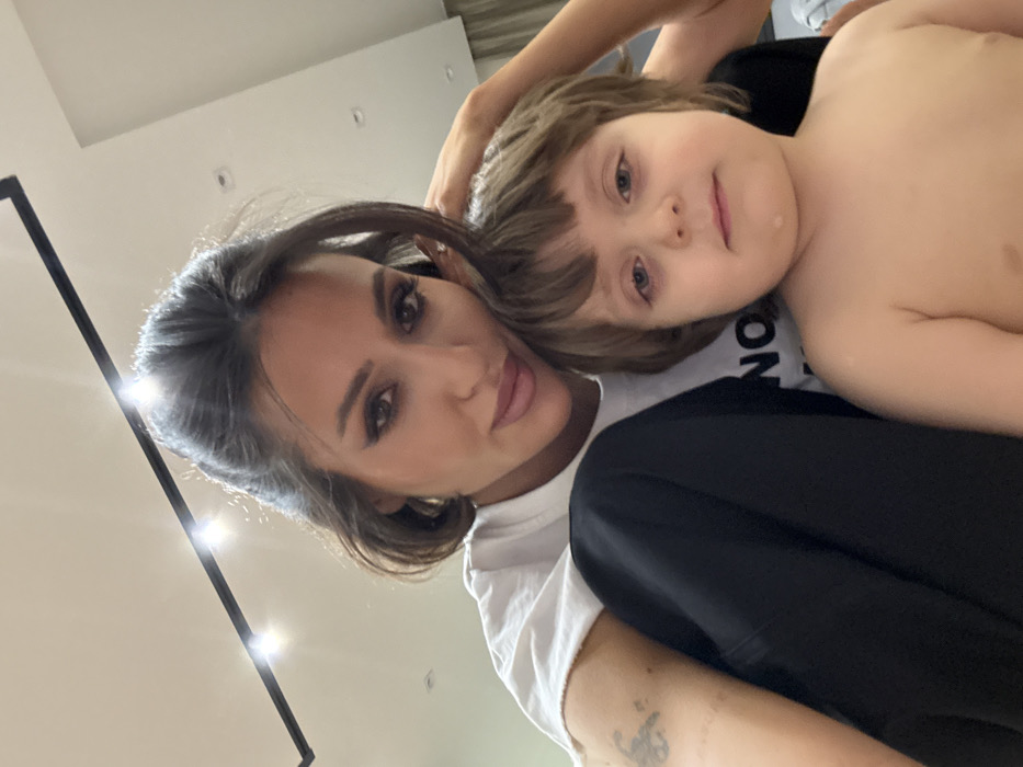 A woman and a young child are closely posed in a selfie. The woman has light skin, dark hair, and wears a white t-shirt. The child, who appears younger, is partially shirtless with light hair and rests his head against her. They both look directly at the camera. The background is bright, with a glimpse of modern lighting fixtures and what looks like a portion of a doorframe or panel.