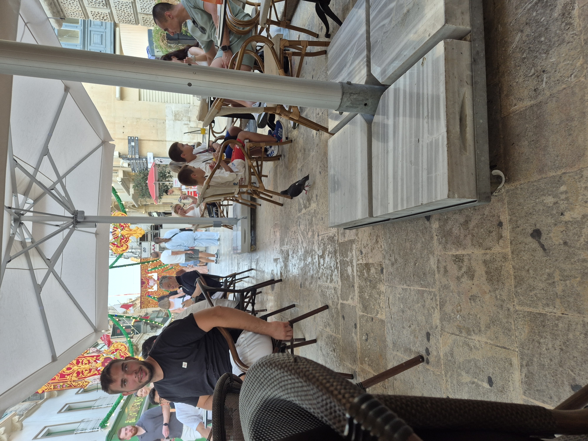 The image captures an outdoor seating area at a cafe or restaurant in Valletta, Malta, under a large white umbrella. A young man in a black t-shirt and white shorts is seated at a table in the foreground, looking towards the camera with a slight smile. He is partially obscured by a wicker chair. In the background, several other people are visible, seated at tables and walking around. The setting appears to be a pedestrian street with buildings and festive decorations, suggesting a public event or celebration. A pigeon is seen walking on the stone-tiled ground in the mid-ground. The overall ambiance is casual and lively, indicative of a typical day in a busy European city.