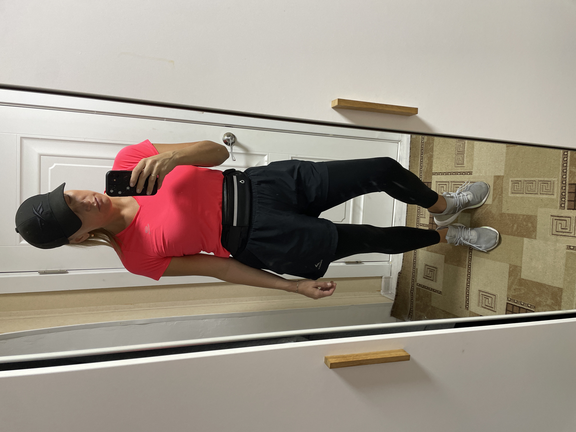 A woman in athletic wear takes a selfie in a mirror. She is wearing a pink t-shirt, black leggings, black athletic shorts, grey sneakers, and a black baseball cap. She is holding a black smartphone in her left hand. The mirror reflects a door, a carpet with geometric patterns, and a white cabinet with wooden handles. The location is not identifiable as Baranovka, Russia.