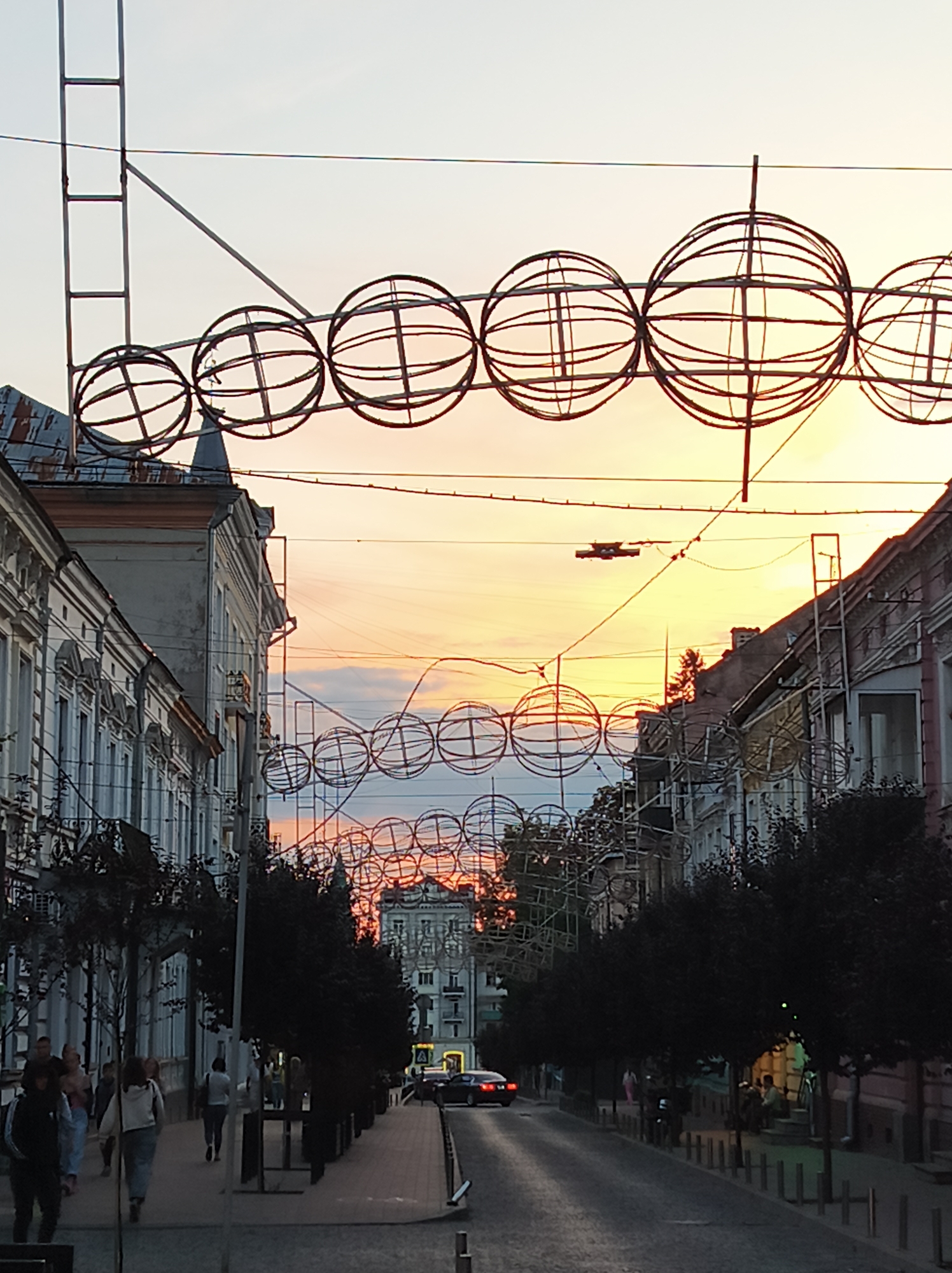 The image depicts a street scene in Ternopil, Ukraine, during what appears to be sunset or dusk. The sky is filled with warm hues of orange and yellow, indicating the setting sun. Overhead, large, spherical wireframe decorations are strung across the street, forming an elaborate display. These decorations are suspended by metal structures and wires.
On the left side of the street, a row of multi-story buildings with classical architectural features lines the sidewalk. Some of these buildings show signs of age, with peeling paint and weathered roofs. A few trees with dark foliage are planted along the sidewalk.
On the right side of the street, similar buildings are visible, also with classical architecture. More trees are present, creating a shaded area along the sidewalk.
A paved road runs down the center of the street, with a parked car visible in the distance. Several people are walking along the sidewalks on both sides of the street, appearing to be engaged in casual activity. The overall atmosphere suggests a public space in an urban environment, possibly a commercial or residential area decorated for an event or season. The streetlights are not yet illuminated, reinforcing the impression of twilight.