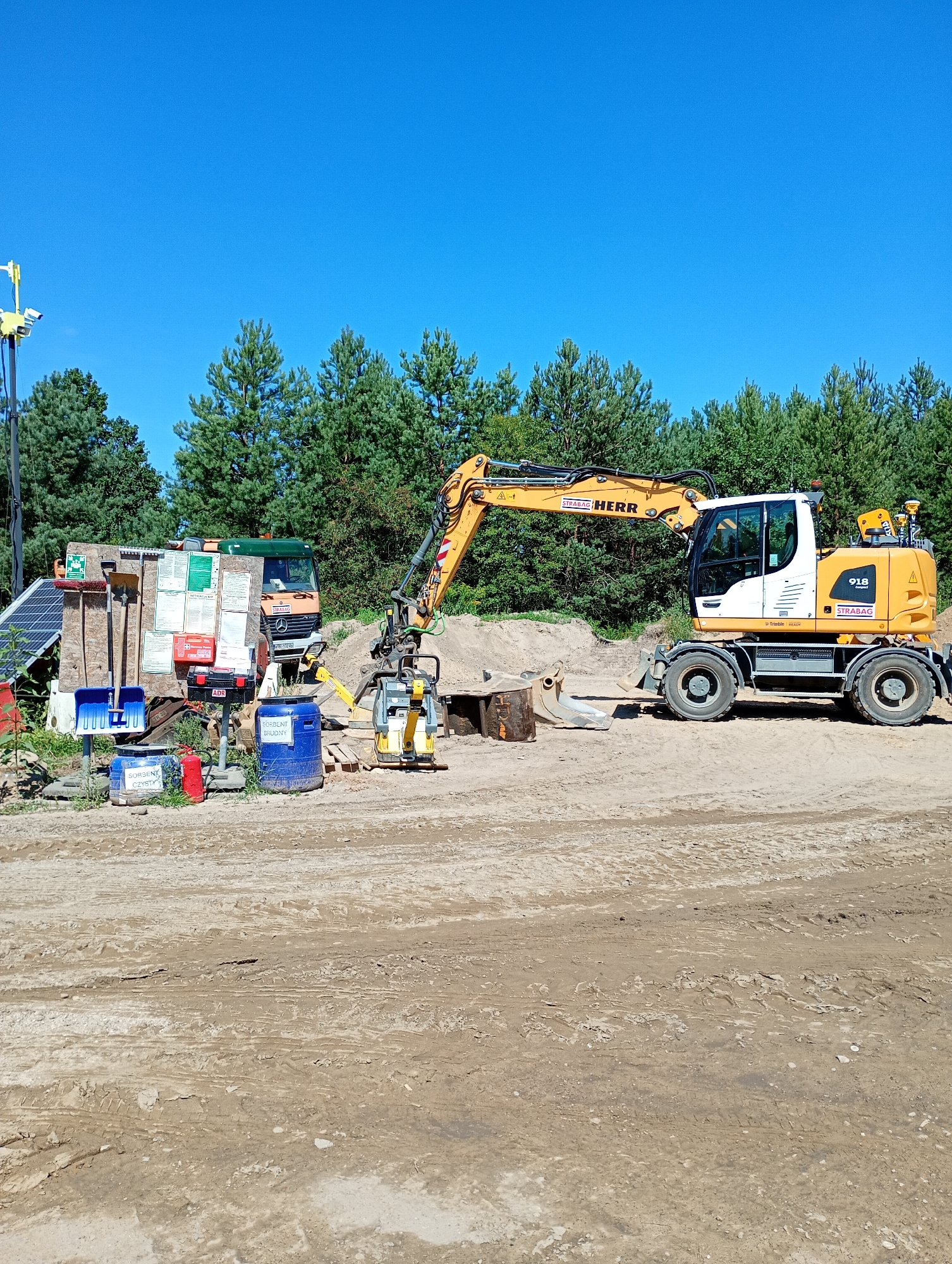 The image depicts an outdoor construction site on a clear, sunny day with a bright blue sky. The terrain is sandy and dusty, with visible tire tracks from heavy machinery. In the background, a dense line of green pine trees forms the edge of the site.

Dominating the scene is a large yellow and white excavator with the brand name "STRABAG HERR" visible on its boom. The excavator is parked on the dirt, with its arm partially extended. Adjacent to the excavator, a smaller, yellow compactor machine is also present.

To the left of the excavator, a collection of items is organized on a wooden board attached to a stand. These items include various tools such as brooms and shovels, a first-aid kit, and several safety and informational signs. Nearby are two blue barrels, one labeled "SORBENT CZYSZCZACY" (cleaning sorbent) and another with the same label. A red fire extinguisher stands beside them. To the far left, a solar panel array is visible, connected to a pole with cameras and lights. Behind the tools and barrels, part of a green and orange truck is visible.

The scene appears to be within or adjacent to the city of Grabówka, Poland, indicated by the presence of construction equipment and signage, commonly found at such locations. No people are visible in the image. The overall impression is that of an active construction or roadwork site.