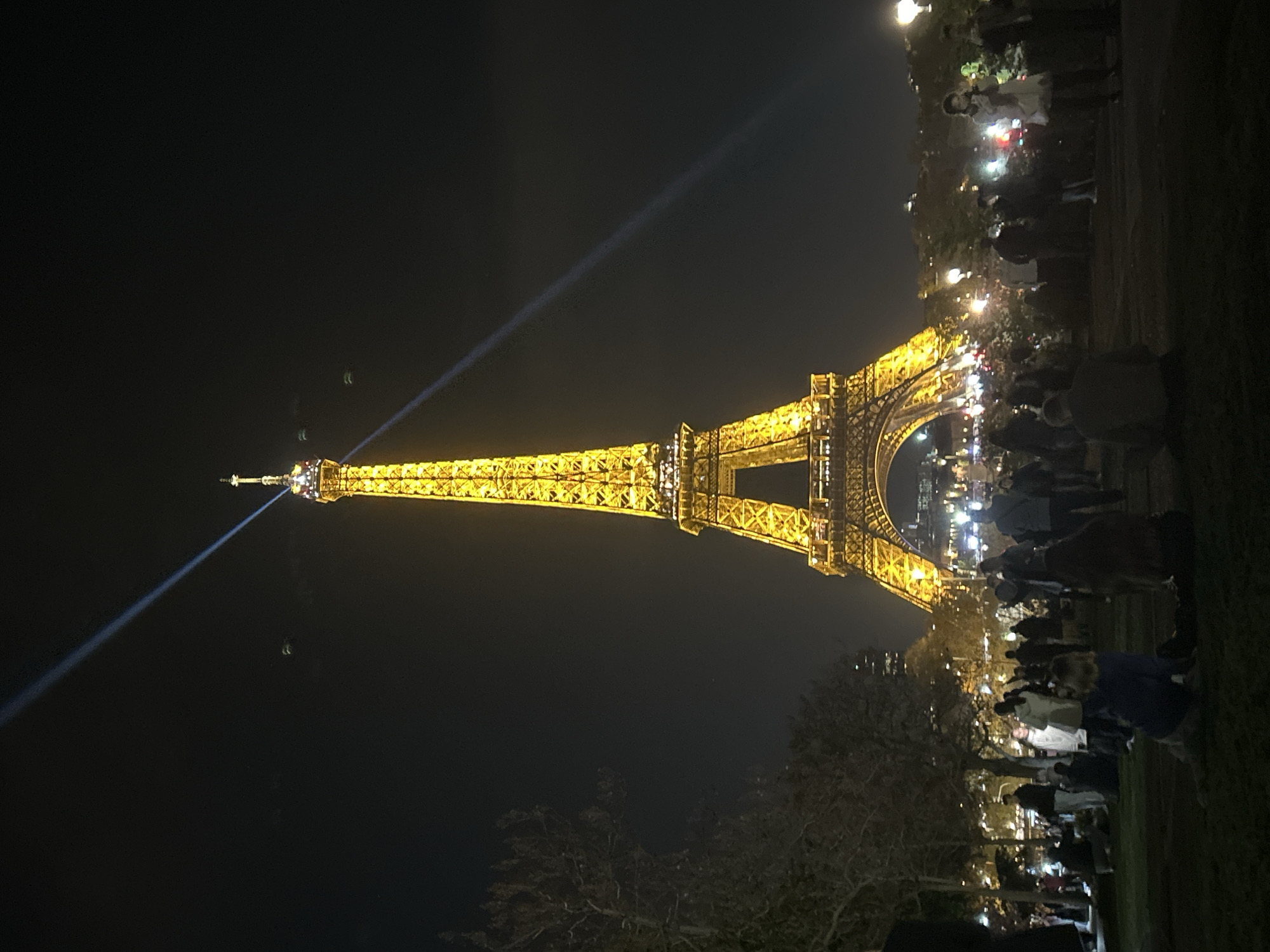 The Eiffel Tower in Paris, France, is illuminated at night. A bright beam of light emanates from the top of the tower. A crowd of people is gathered at the base, partially obscured by trees, with some holding up phones. The sky is dark. The image is taken at night.