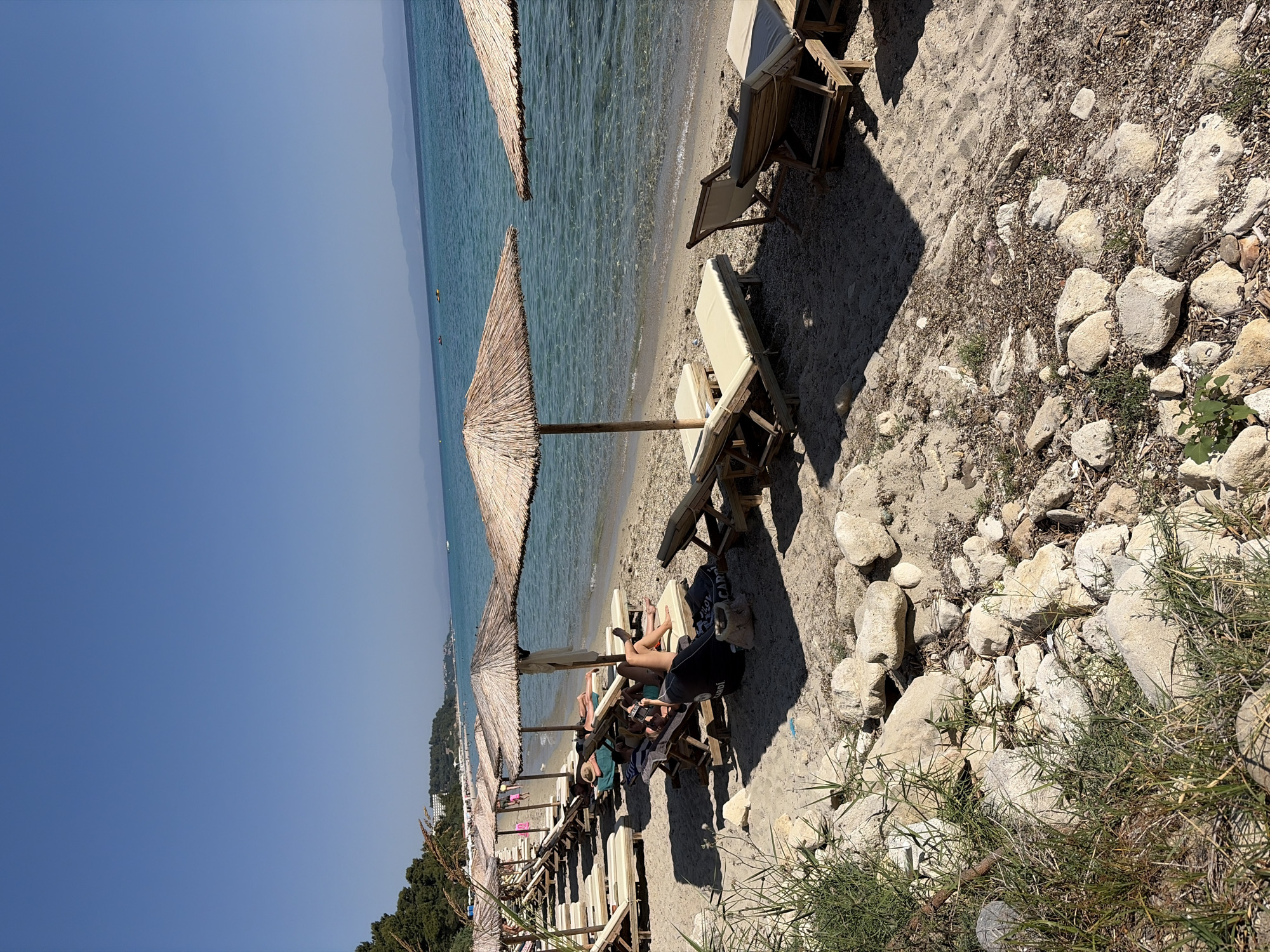 This image captures a sunny day at a beach in Kassandra, Greece. The scene features a row of lounge chairs and thatched umbrellas on a sandy and rocky shore. The clear turquoise water of the sea gently laps the beach.
Several people are relaxing on the lounge chairs under the umbrellas. One person in the foreground is wearing black shorts and a black top, with their legs stretched out. Another person, wearing a straw hat, is also visible on a lounge chair. The atmosphere appears relaxed and tranquil, typical of a summer vacation.
The time of day is clearly daytime, indicated by the bright sunlight casting distinct shadows from the umbrellas and lounge chairs onto the sand. The weather seems to be warm and clear, with a deep blue sky dominating the upper portion of the image. The presence of pine trees and lush greenery in the background suggests a Mediterranean coastal setting. The beach is dotted with a variety of rocks, from small pebbles to larger stones, adding to the natural landscape. In the distance, a hazy outline of hills or mountains can be seen across the water. The image is oriented vertically. No visible text is present.