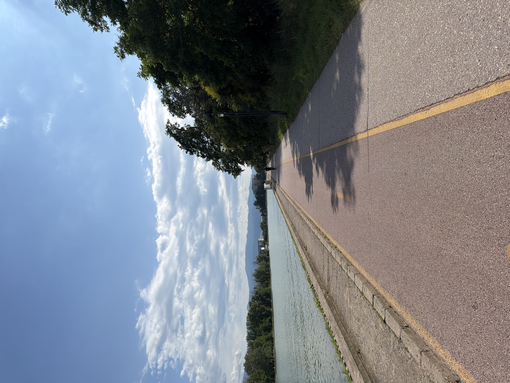 The image depicts a wide, paved recreational path running alongside a body of water, likely a lake or river, with lush green trees lining the opposite bank. The path is divided by a yellow line, with one side appearing to be a red-tinted asphalt and the other a grey asphalt, suggesting distinct lanes for different activities. Several people are visible on the path; one individual is on a bicycle in the distance, and another appears to be walking. The water is calm, reflecting the blue sky dotted with white, fluffy clouds. In the background, a line of trees and distant hills or mountains can be seen. This scene is consistent with the rowing canal in Plovdiv, Bulgaria, a popular area for recreation and sports. The lighting suggests it is daytime, likely late morning or afternoon given the presence of shadows.