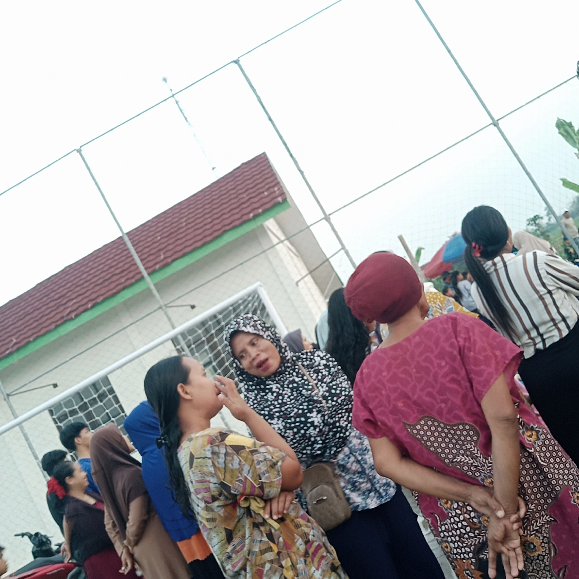 In the city of Tegowanu Wetan, Indonesia, a group of people are gathered outdoors, likely at a sporting field given the presence of a net and a building with a reddish-brown tiled roof and green trim. In the foreground, a woman wearing a patterned dress with yellow, brown, and pink hues is facing left and touching her face. To her right, another woman in a black and white spotted hijab is looking towards the woman in the patterned dress, with her mouth open as if speaking. Further to the right, a woman in a maroon headscarf and a magenta batik-patterned dress is seen from the back, with her hands clasped behind her. In the mid-ground, other individuals are visible, including a person in a striped shirt and a blue hijab, and a person in a brown hijab. The background shows more people, some foliage, and a lighter sky. The overall atmosphere suggests a social gathering or event.