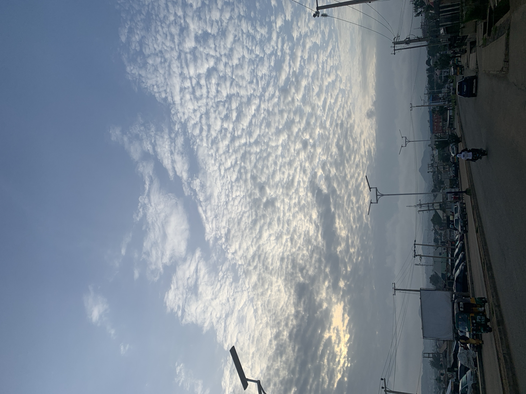 The image depicts a street scene in Rafin Par, Nigeria, under a vast sky filled with altocumulus clouds. The sky is a dominant feature, with a clear blue expanse overhead transitioning to a dense pattern of small, fluffy clouds that form a textured layer across the sky. The lighting suggests either early morning or late afternoon, with a soft, diffused glow.
In the foreground, a paved road runs through the scene, with vehicles and a pedestrian walkway visible. A distinctive green and yellow motorized tricycle, often used as a taxi, is parked on the side of the road. Several cars are also parked or driving along the street. A large, blank white billboard stands prominently on the left side of the road.
Streetlights with modern designs are erected along the road, some with solar panels attached. Power lines are strung between utility poles, crisscrossing the upper portion of the scene. In the background, buildings of various sizes and styles form a townscape, with hints of residential and commercial structures. Beyond the town, rolling hills or mountains can be seen in the distance, softened by the atmospheric conditions. The overall impression is one of a typical urban environment in Nigeria, with a focus on the sky's dramatic cloud formations.