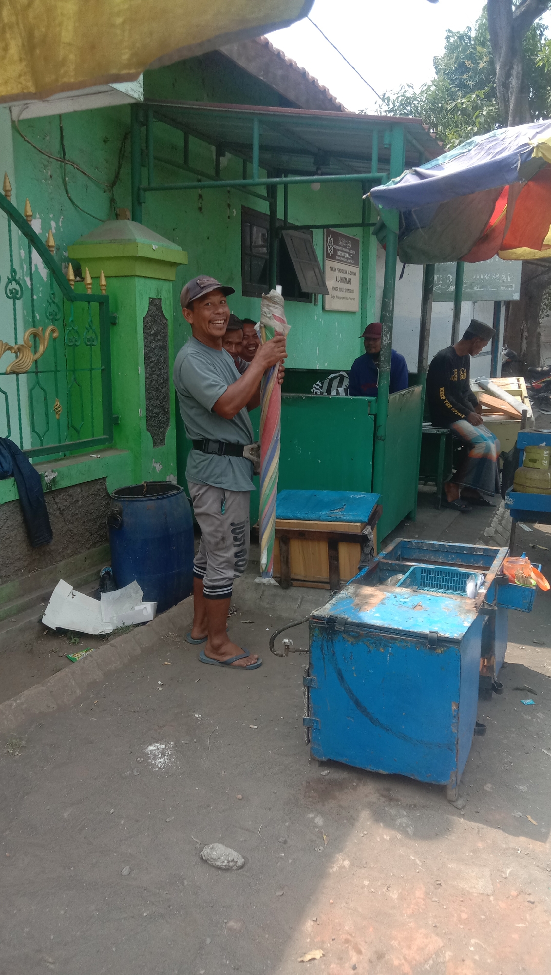 In Karang Anyar, Indonesia, a man in a grey t-shirt and cap stands facing forward, holding a rolled-up umbrella with colorful stripes. He is smiling and appears to be a street vendor. Behind him, two other men are partially visible. The setting is outdoors, with a green building that has an open window and a sign with Indonesian text. To the right of the man, a blue, weathered metal cart with a lid sits on the ground, possibly used for selling goods. In the background, there's a brown wooden box with a blue lid, and a green structure with a roof. The ground is paved with asphalt. The scene suggests a typical day in a busy street market or a small business area.