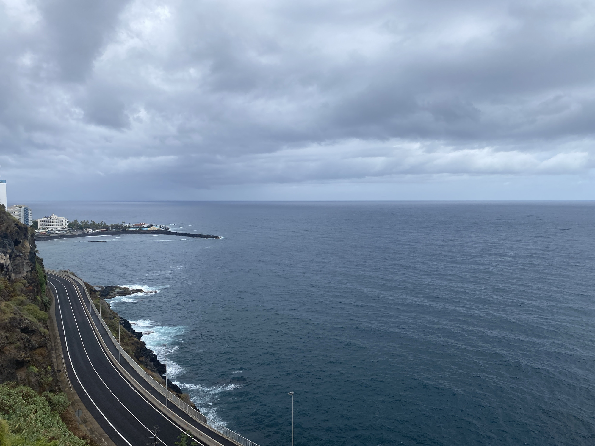 This is a high-angle, full shot of a coastal road winding along a cliff face overlooking the ocean under a cloudy sky. The road is asphalt with distinct white lane markings and is bordered by a metal guardrail. Lush green vegetation grows on the left side of the road, clinging to the rocky cliff. The ocean is a deep blue, with white waves crashing against the rocky shore below. In the distance, a town with white buildings and a pier can be seen. The overall scene conveys a sense of dramatic natural beauty and modern infrastructure coexisting. The location appears to be the coastal road leading towards the town of Puerto de la Cruz in Tenerife, Spain, known for its dramatic cliffs and ocean vistas. There are no people visible in the image.