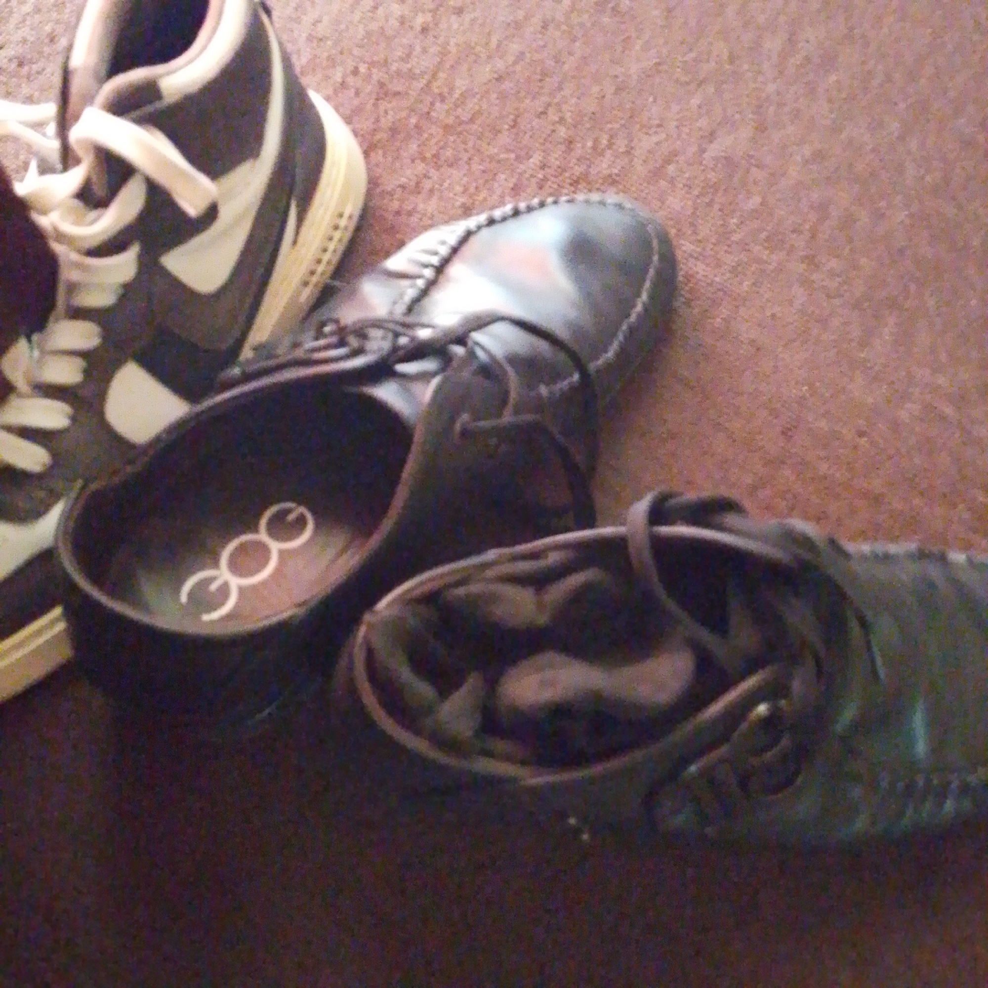 The image shows a close-up, overhead view of three pairs of shoes on a carpeted surface. In the upper left is a black and white Nike sneaker, with its laces visible. To the right of the sneaker is a black leather loafer with visible stitching and laces. In the foreground, to the right, is another black leather shoe, possibly a loafer or moccasin, with visible stitching. In the center of the image is a close-up of the interior of a black shoe, revealing a white logo with three interconnected circles. The carpet appears to be a low-pile, brown or reddish-brown color, consistent with flooring found in residential or commercial spaces in Jalingo, Nigeria. No people or activities are depicted in the image.
