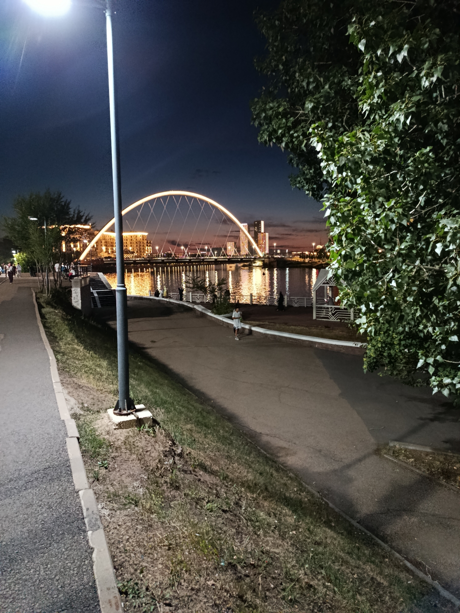 The image depicts a nighttime scene in Astana, Kazakhstan, specifically along the Ishim River embankment. In the foreground, a paved walkway runs alongside a grassy area. A tall lamppost stands prominently in the left-center of the frame, its light illuminating the immediate surroundings. Further into the background, a modern, illuminated arched bridge spans the river, with several tall buildings visible on the opposite bank, their lights reflecting on the water. The sky is dark, indicating it is nighttime. A person is visible walking on the walkway, and other individuals are present on the embankment near the river. Lush green trees are present on the right side of the image, partially obscuring the view of the distant cityscape. The overall atmosphere is serene and urban.