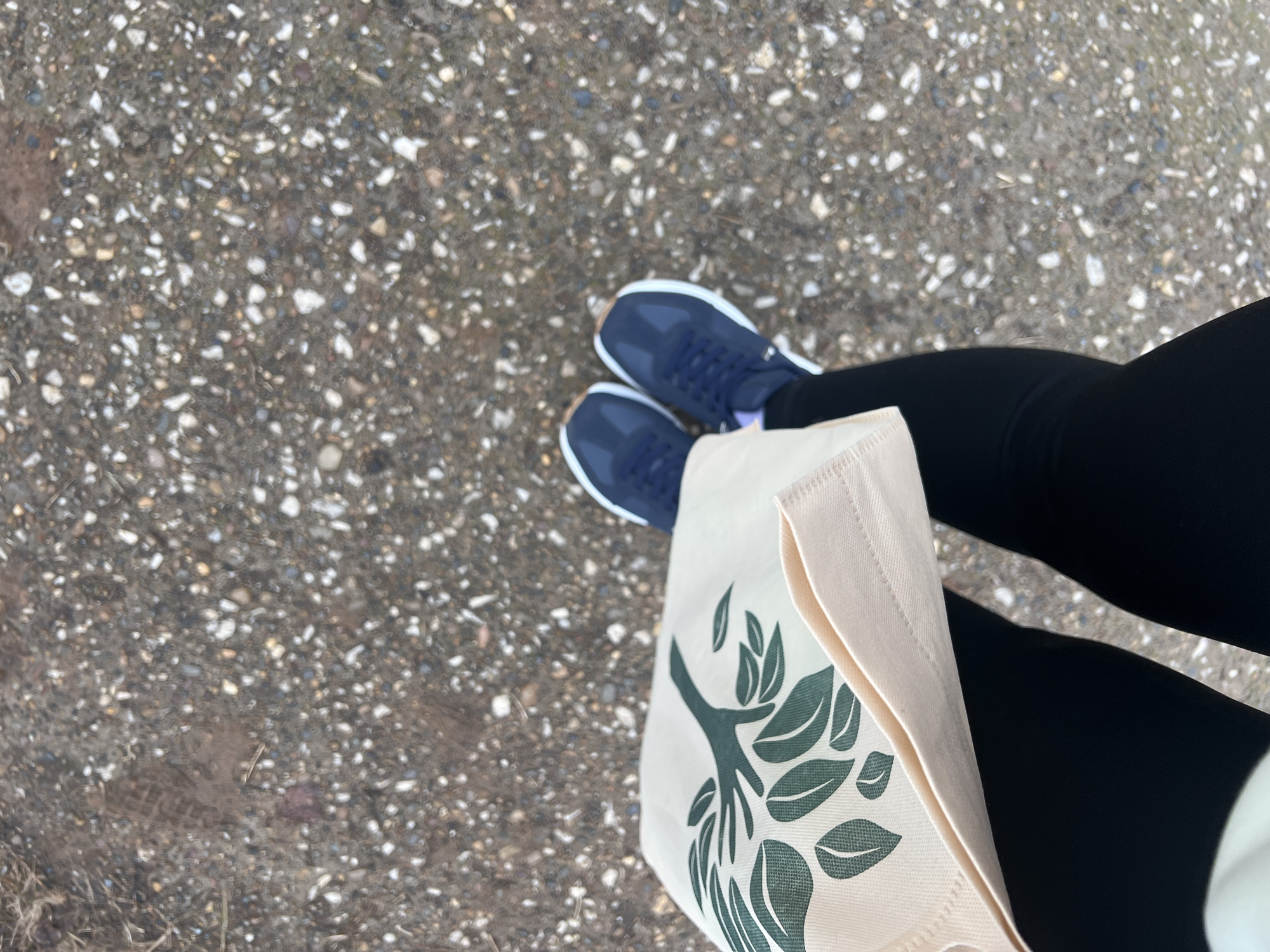 The image shows a person's legs, feet, and part of a tote bag. The person is wearing black leggings and navy blue sneakers with white soles. A light-colored tote bag with a green botanical print is positioned beside their legs. The ground is paved with speckled asphalt. The image's location is unavailable.