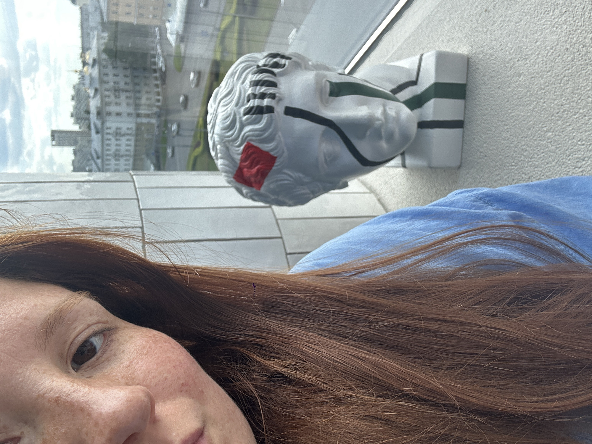This close-up, rotated selfie features a woman and a decorative bust positioned indoors, overlooking a city through a large window. The woman occupies the lower left portion of the frame, with her reddish-brown hair flowing across the bottom. Her visible features include her left eye, which is dark brown, and a cheek and nose area covered in freckles. She is wearing a light blue garment. Her expression appears neutral.
To her right, slightly above her head in the rotated image, is a white bust resembling a classical Roman or Greek head, characterized by curly hair details. This bust is adorned with modern, abstract design elements: black stripes running across the top of the head and eye area, and green lines extending from the back of the head down its base. A distinct red, textured square is painted on the side of the bust's head. The bust rests on a square base with matching black and green lines.
Through the window in the background, a cityscape is visible, suggesting a daytime setting with a partly cloudy sky. Numerous buildings of varying architectural styles, some appearing older, line streets where cars can be seen. The reflections on the glass obscure some details of the urban environment. The overall scene suggests a moment captured from an elevated position within a city like Nizhny Novgorod, Russia, focusing on a personal interaction with art against an urban backdrop. There is no visible text in the image.