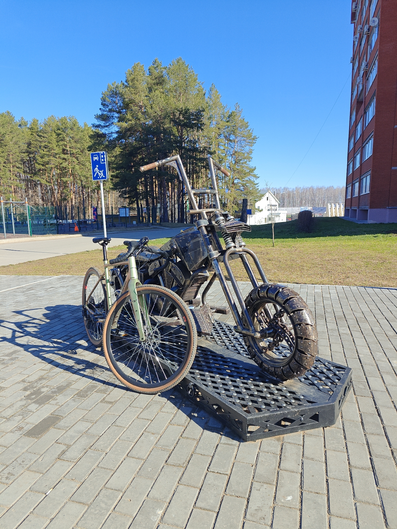 The image captures a sunny day in Chaikovsky, Russia, where a metal sculpture of a motorcycle is prominently displayed on a black, grated platform set on a brick-paved area. A bicycle is parked beside the sculpture. In the background, there is a building with red brick, a patch of green grass, trees, and a blue pedestrian and parking sign. The sky is clear and blue.