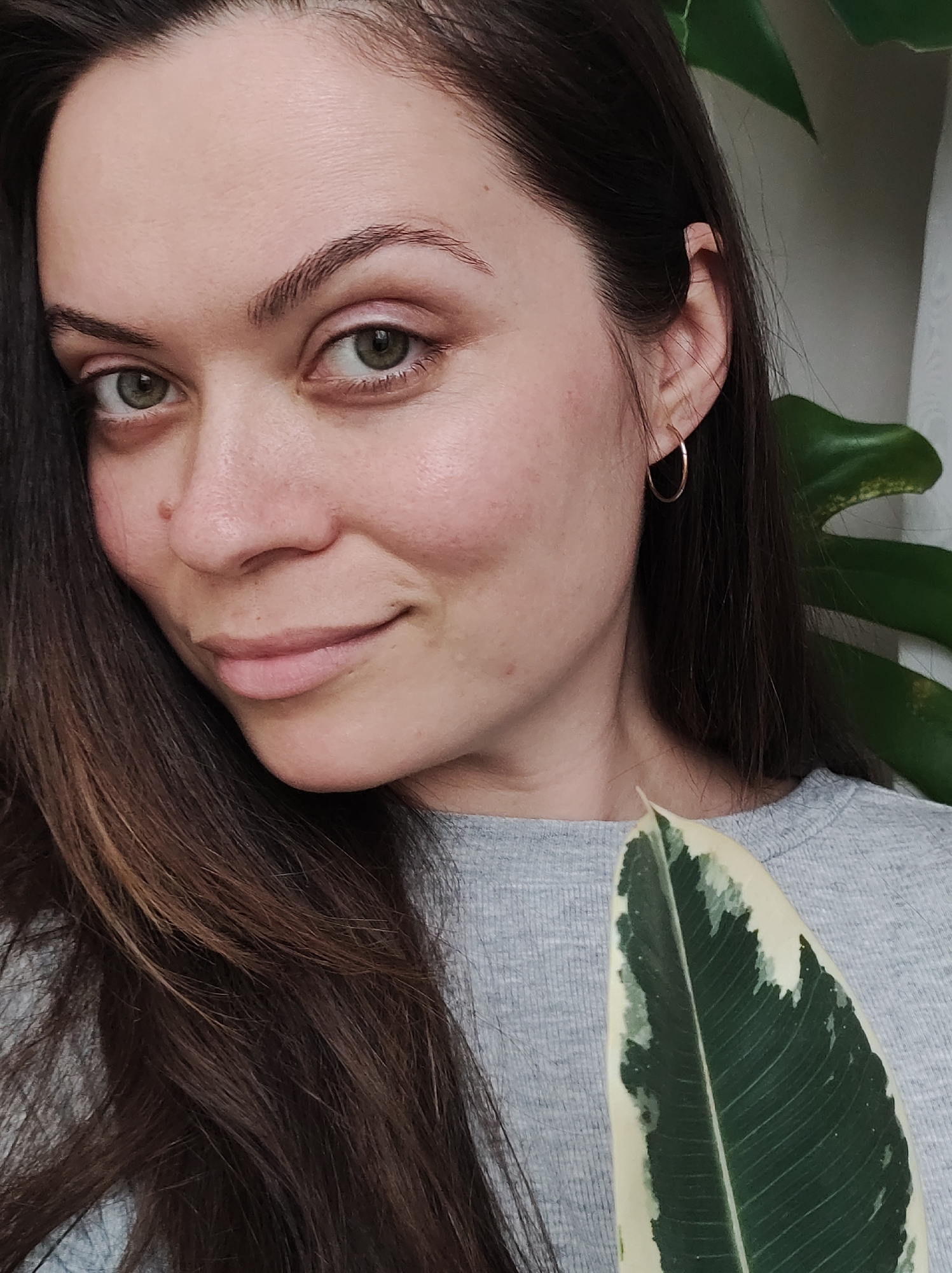 A woman with long brown hair, green eyes, and light skin is posing for a close-up selfie. She is wearing a gray t-shirt and gold hoop earrings. Her expression is pleasant, with a slight smile. In the lower right corner of the frame, she is holding a large green and white variegated leaf. Behind her, there is a larger green plant, partially visible, and a white curtain. There is no information to suggest the location is in the city of Chaikovsky, Russia.