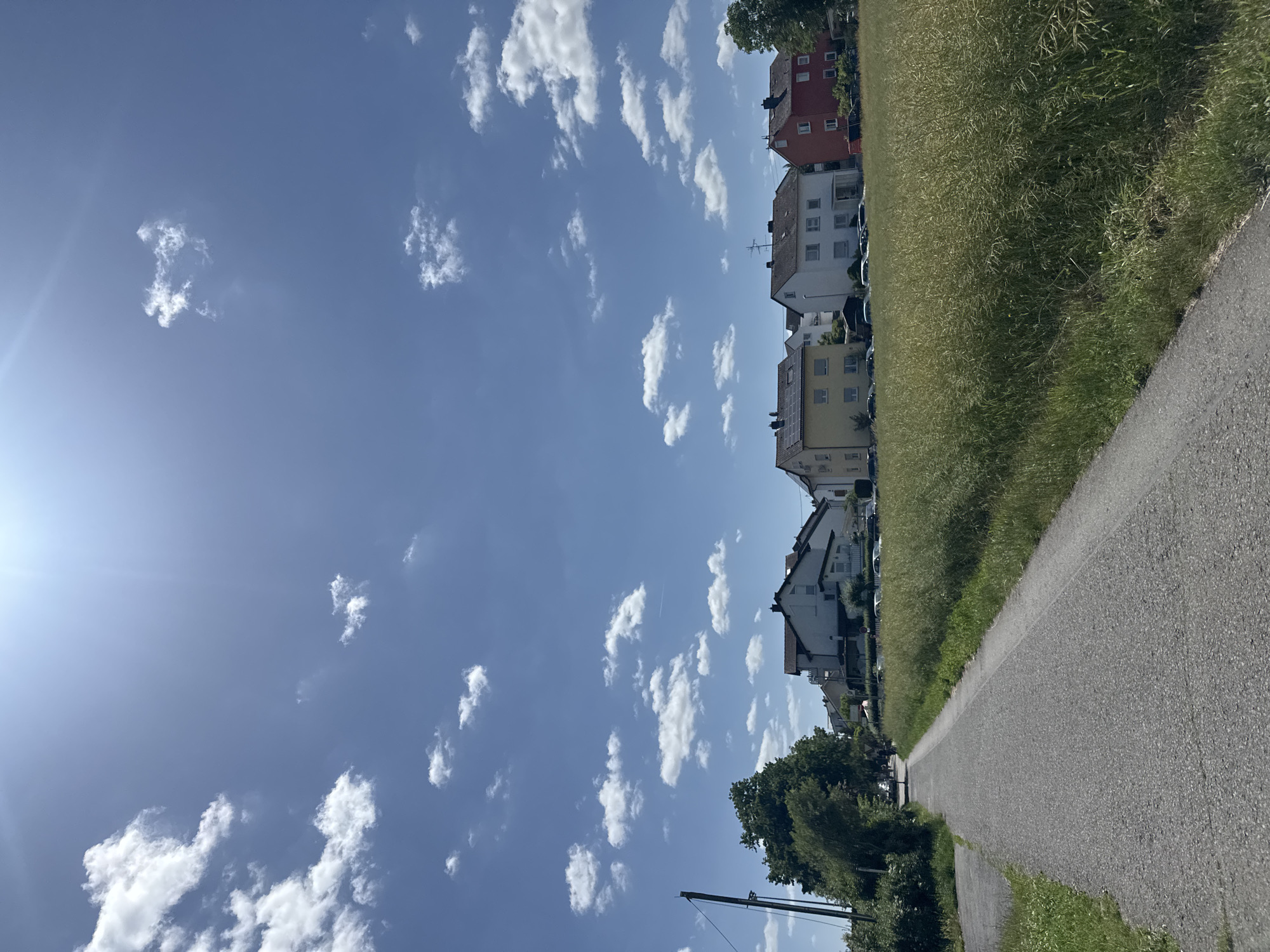 The image depicts a bright, sunlit daytime scene in Hattersheim am Main, Germany. The sky, occupying the upper two-thirds of the frame, is a clear blue adorned with scattered white cumulus clouds. A strong sun glare and lens flare emanate from the upper right. A paved asphalt path, likely for pedestrians or cyclists, traverses the lower left foreground, curving gently left and receding into the distance, bordered by green grass. To the right of the path lies a wide field of tall, light green-yellowish crops. In the midground, a row of residential houses is visible: a yellow two-story house with a dark roof fitted with solar panels, followed by a white two-story house with a dark roof and a small covered entrance, and then a red two-story house with a dark roof. Each house features multiple windows. Several parked vehicles are partially visible along the street in front of the houses. The left side of the path is lined with dense green trees and a dark utility pole. In the far distance on the path, a few indistinct human figures are discernible.