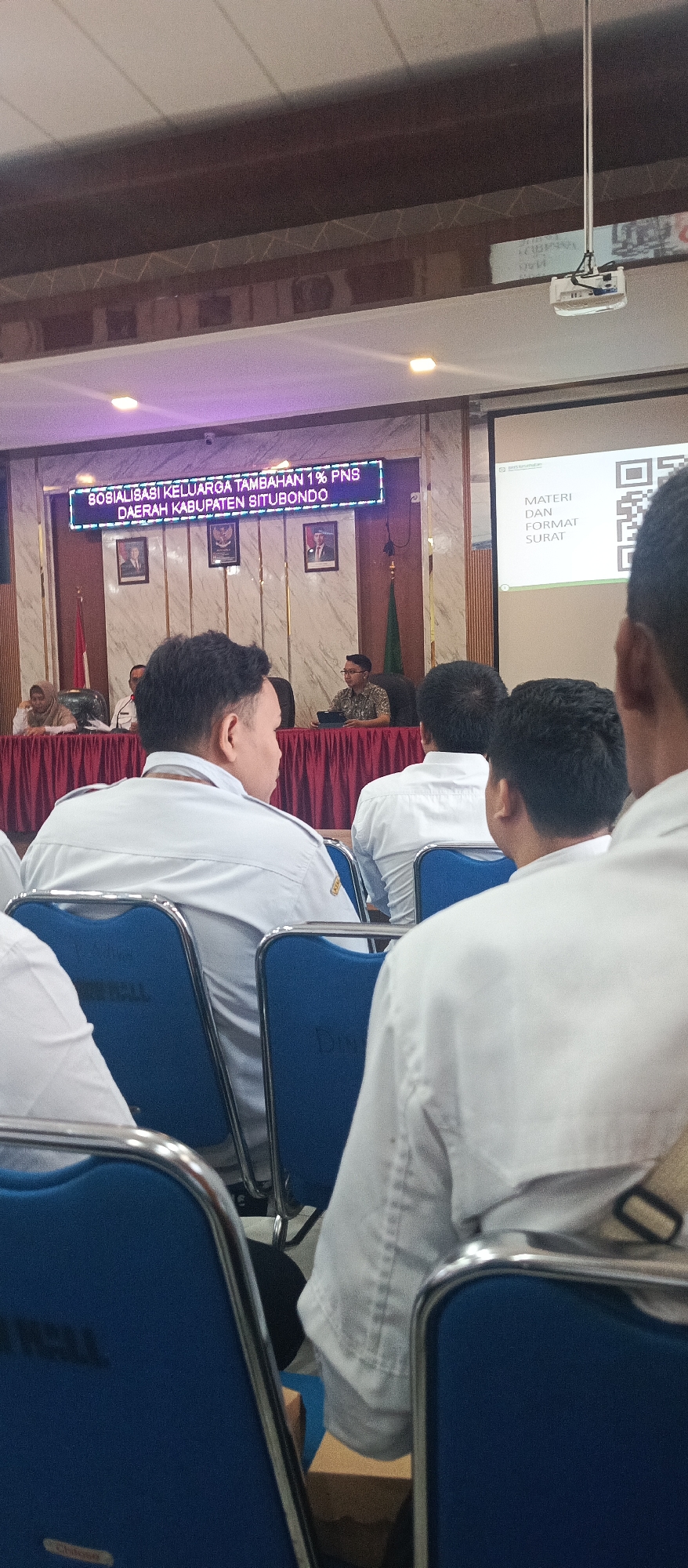 The image depicts an indoor meeting or presentation in Situbondo, Indonesia. In the foreground, the backs of several men in white shirts are visible, seated in blue chairs with silver frames. The chairs are arranged in rows facing a stage or presentation area.
The stage area features a large digital screen displaying text in Indonesian: "SOSIALISASI KELUARGA TAMBAHAN 1% PNS DAERAH KABUPATEN SITUBONDO," which translates to "Socialization of 1% Additional Family for Civil Servants in Situbondo Regency." Below the screen are framed portraits and a green flag.
To the right of the stage, a projector is mounted on the ceiling. Another screen displays a QR code and the text "MATERI DAN FORMAT SURAT," meaning "Material and Letter Format." A man in a patterned shirt is seated behind a table on the stage, likely the presenter, with a tablet in front of him. Other individuals are seated at tables on the stage. The background is adorned with a deep red curtain. The lighting suggests an organized event.