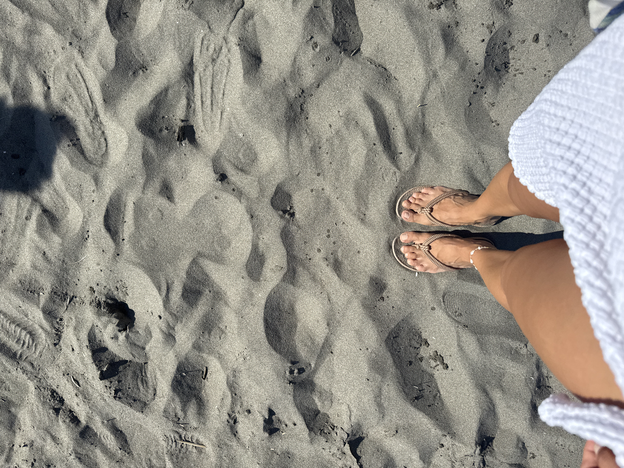 An overhead shot shows a person's feet clad in brown rope flip-flops standing on dark, wet sand. The sand is sculpted with subtle ripples and indentations, likely from the tide or wind. A shadow of the person or an object above them is cast on the sand. The person's legs and a portion of a white, textured towel are visible at the bottom of the frame. The scene appears to be a beach in Donji Štoj, Montenegro, given the dark sand and water's edge proximity often found there.