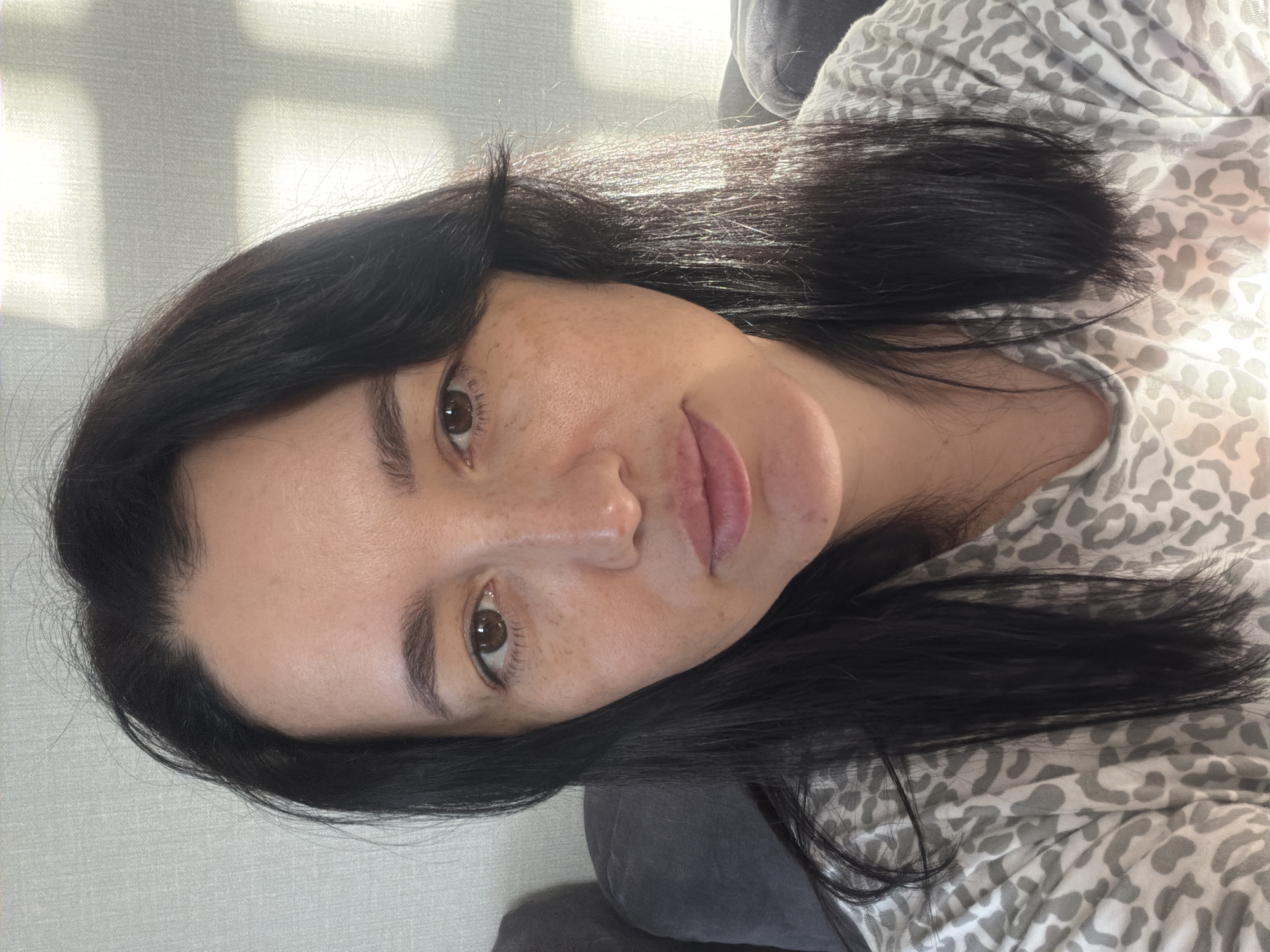 This is a close-up, eye-level selfie of a woman with dark hair and brown eyes, taken indoors. She is wearing a gray and white leopard-print top. Her skin has freckles and appears to be lit by sunlight casting shadows on a light-colored wall behind her. The background also shows a dark gray upholstered surface, possibly a couch. The image does not provide any information about the location in the city of Шмидтово, Russia.